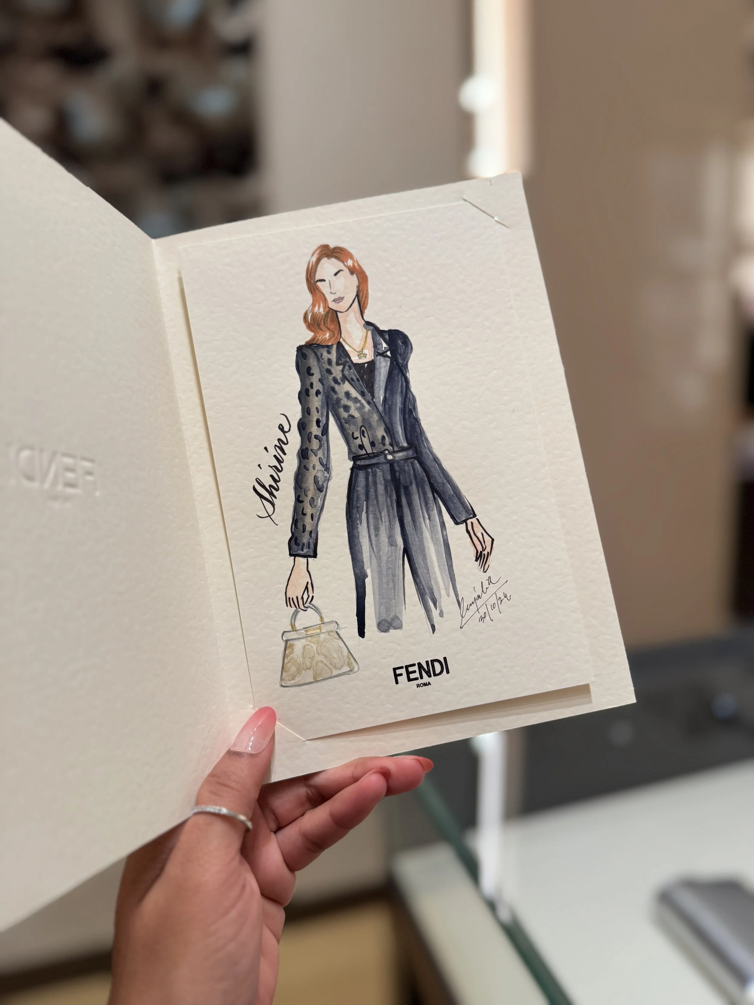 A hand holding a folded fashion illustration card featuring a woman in a leopard print blazer, gray pants, and gold jewelry, with the brand name FENDI at the bottom.