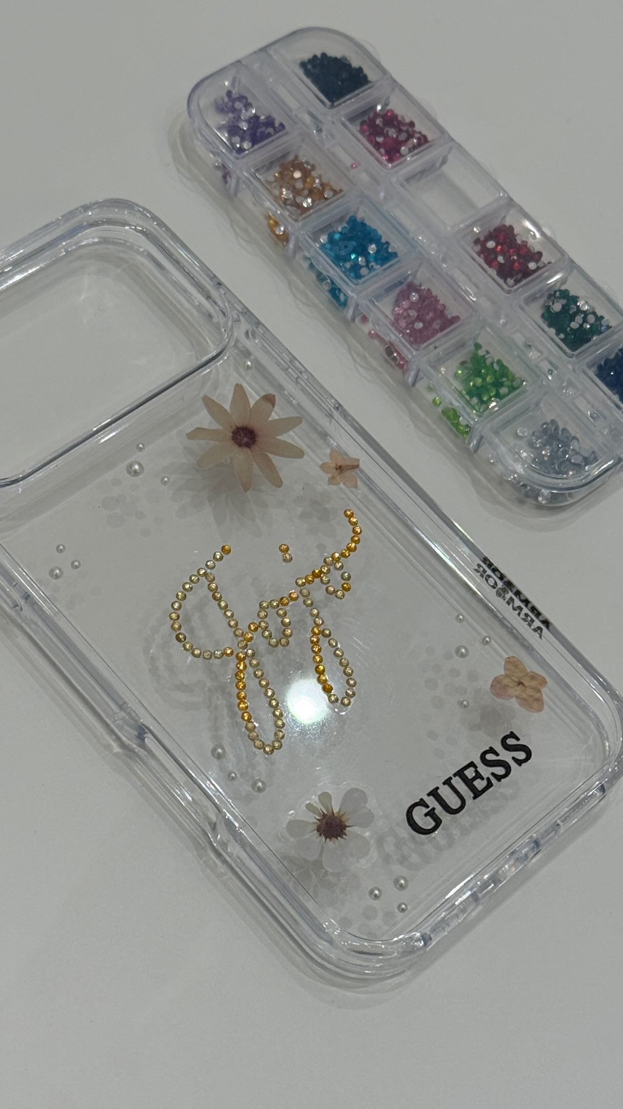 Clear plastic jewelry organizer with gold letter beads spelling 'GEEK' inside, decorated with artificial flowers, and a separate compartment containing small colored rhinestones.