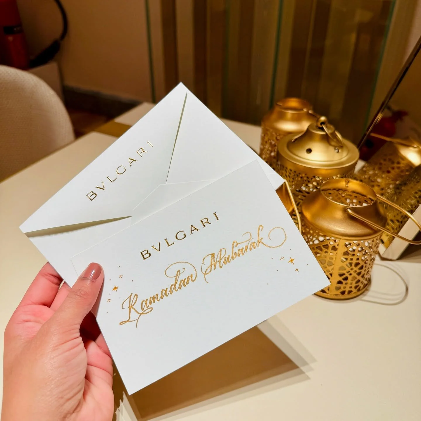 Two white greeting cards with gold lettering and decorations, held by a hand, with gold lanterns on a table in the background.
