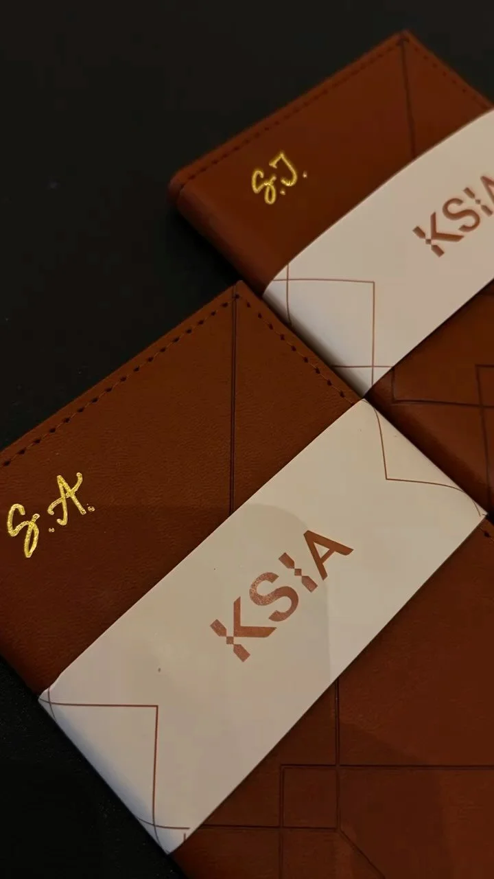 Close-up of two brown leather wallets with gold initials 'S.A.' and white paper bands featuring the red text 'KSI'A' on a dark surface.