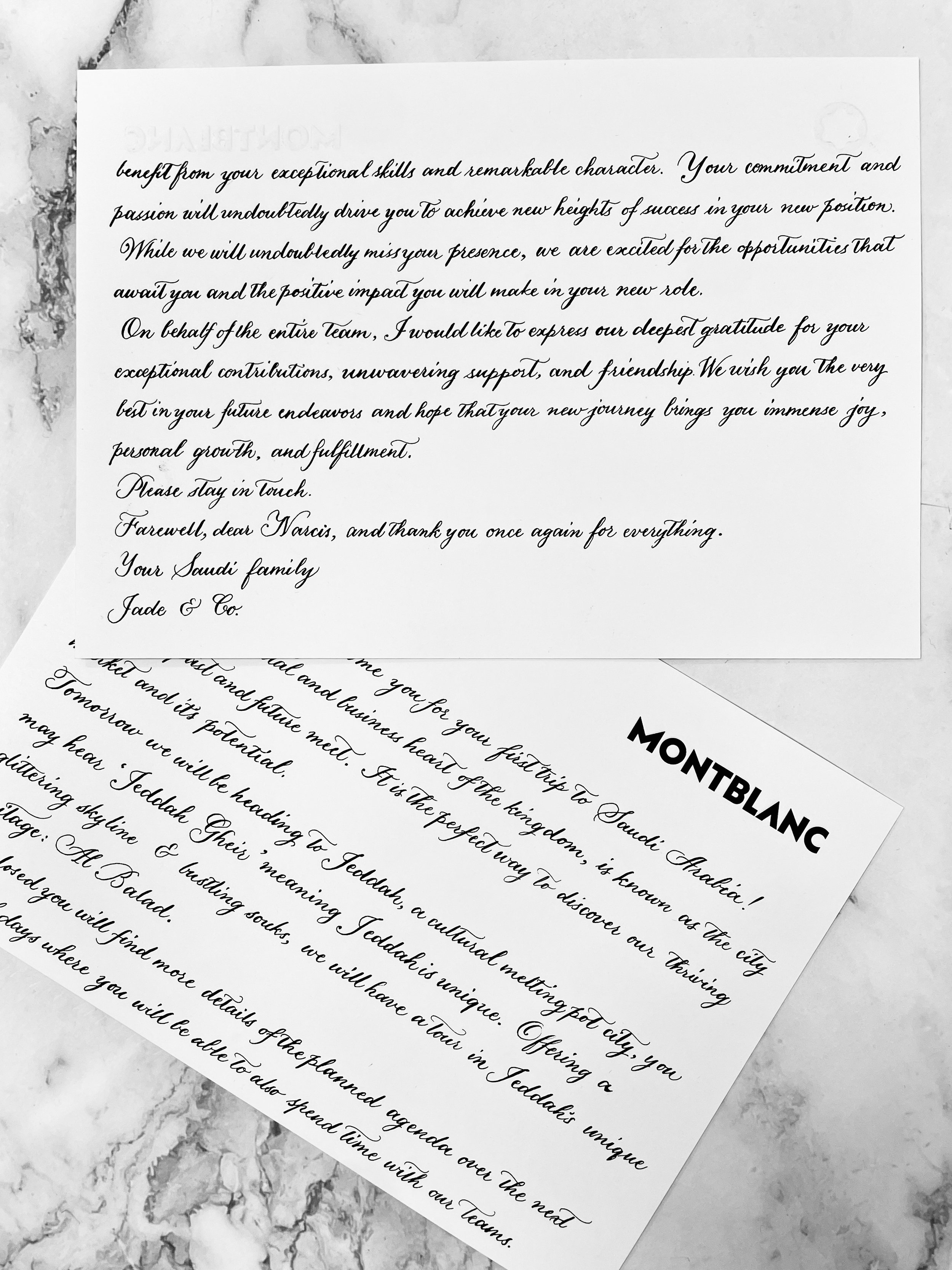 Two sheets of letter-sized paper with handwritten black cursive text on a marble surface. One sheet has the heading 'MONTBLANC' printed in bold, larger font. The handwritten text appears to be a formal letter or note.