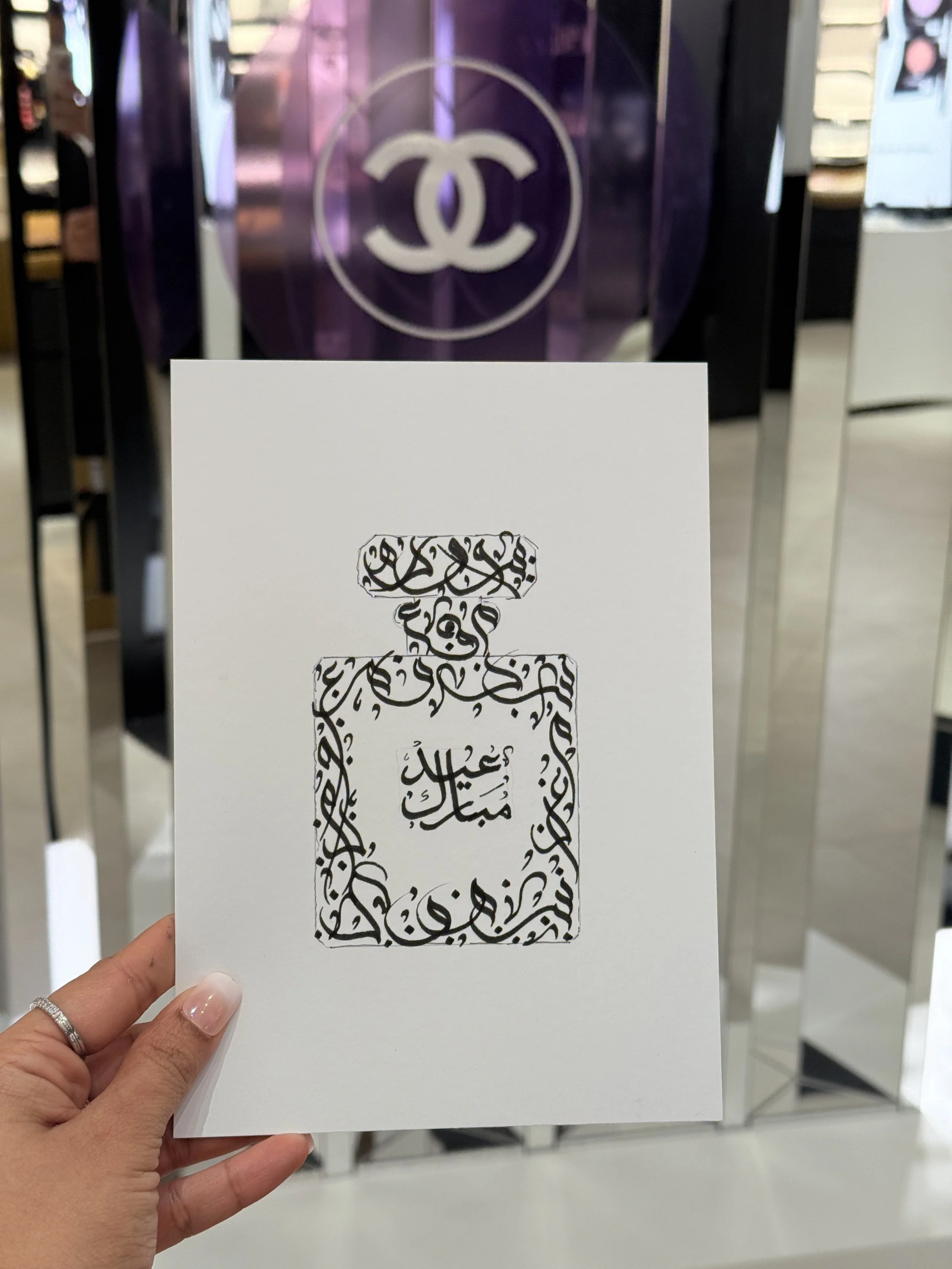A hand holding a white card with black Arabic calligraphy, with a background of a retail store featuring the Chanel logo