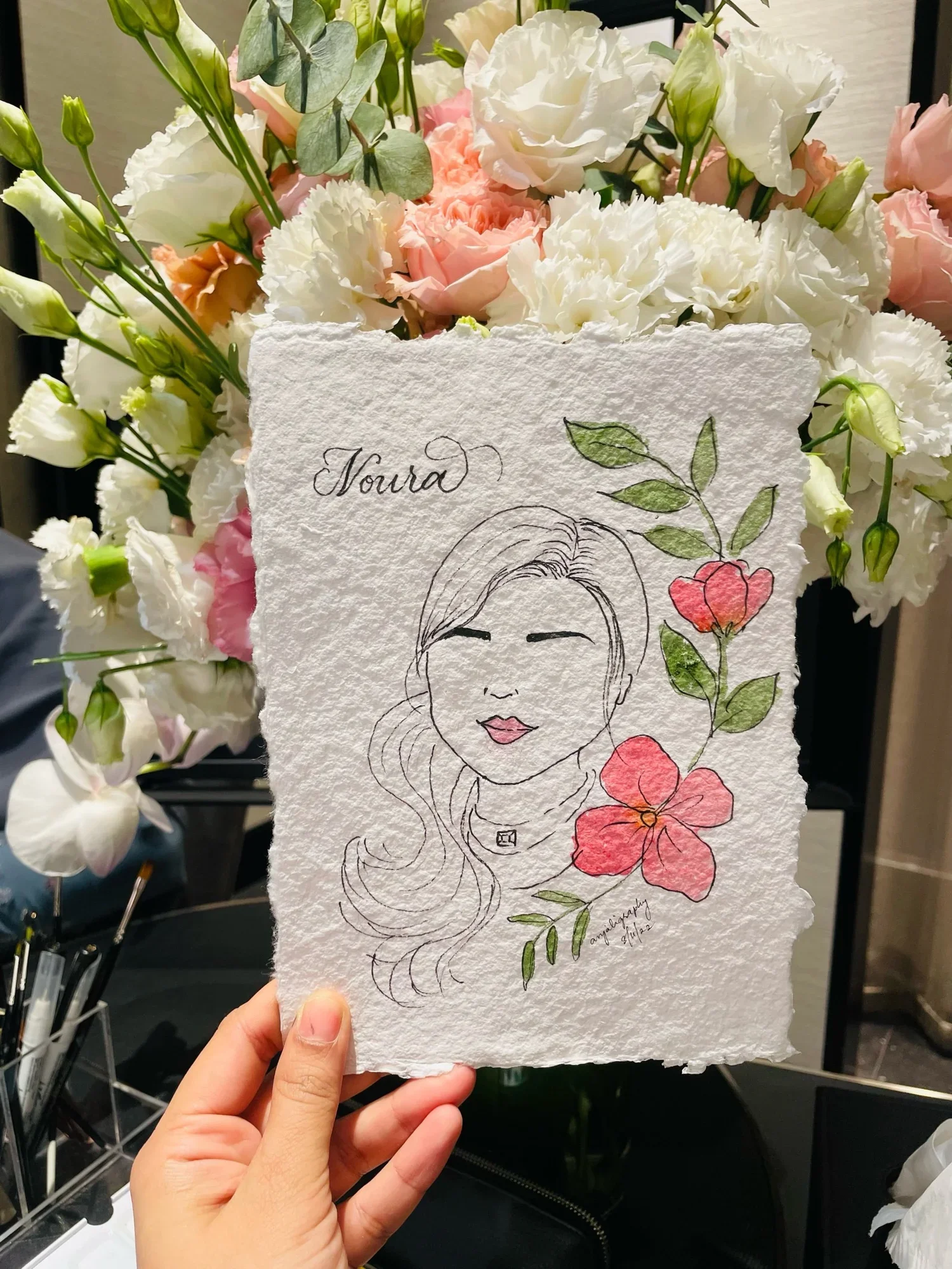 A hand holding a piece of textured white paper with a line drawing of a woman's face, surrounded by colorful flowers and green leaves. In the background, there are arrangements of pink and white flowers.