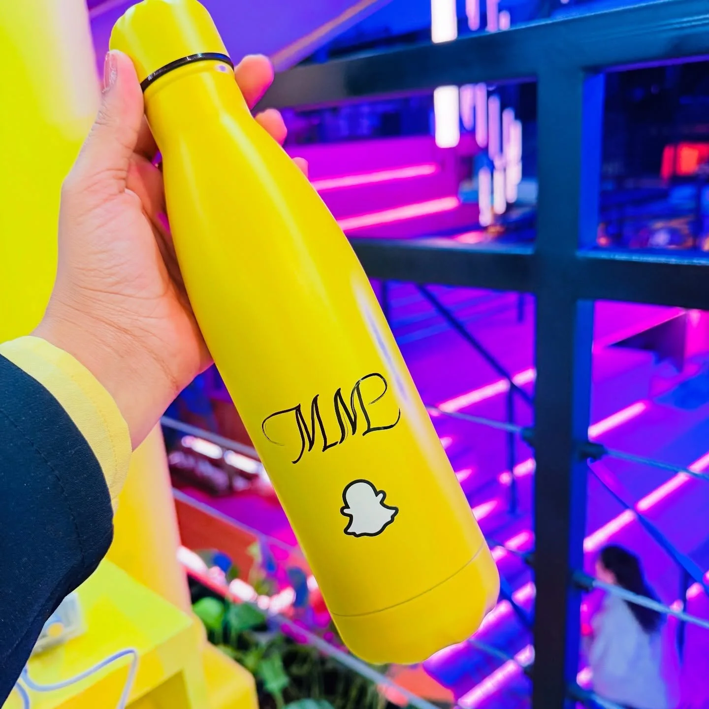 A person holding a yellow bottle with the word 'TUMBL' and the Snapchat ghost logo printed on it, in front of a colorful, neon-lit background.