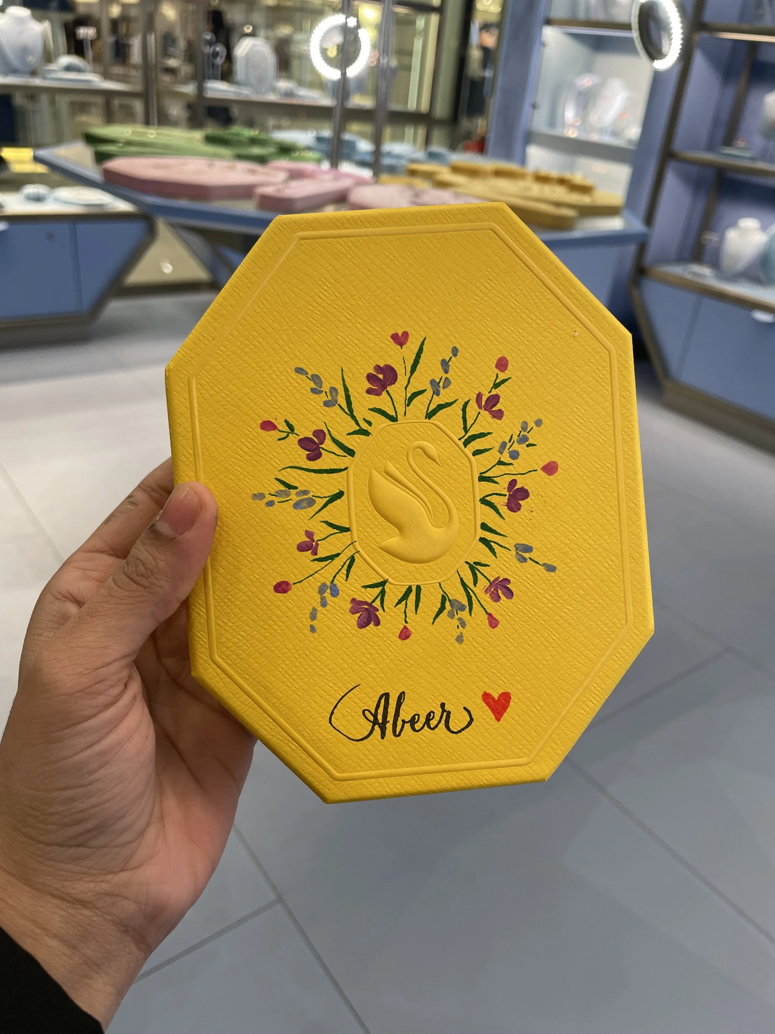Close-up of yellow decorative napkins held in hand, with floral design and the word 'Abeir' written in cursive, in a store with glass shelves and kitchenware in the background.