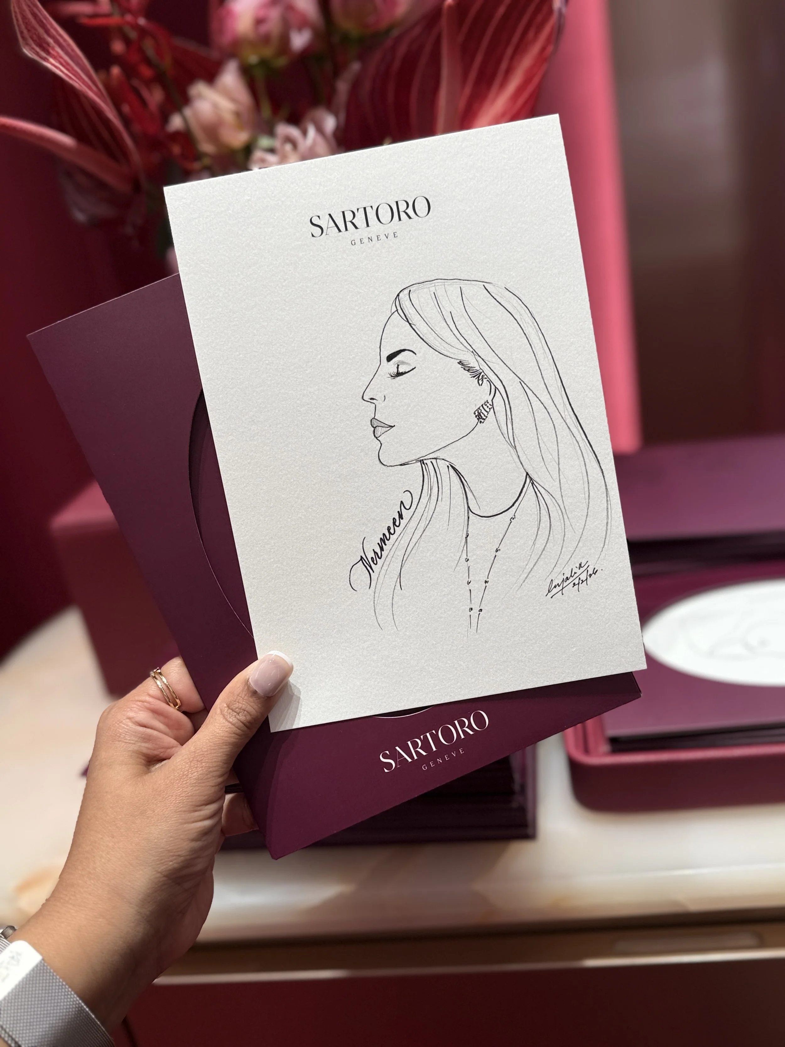 A hand holding a card with a sketch of a woman's profile and the text 'SARTORO GENEVE' at the top, with a pink box filled with flowers in the background.