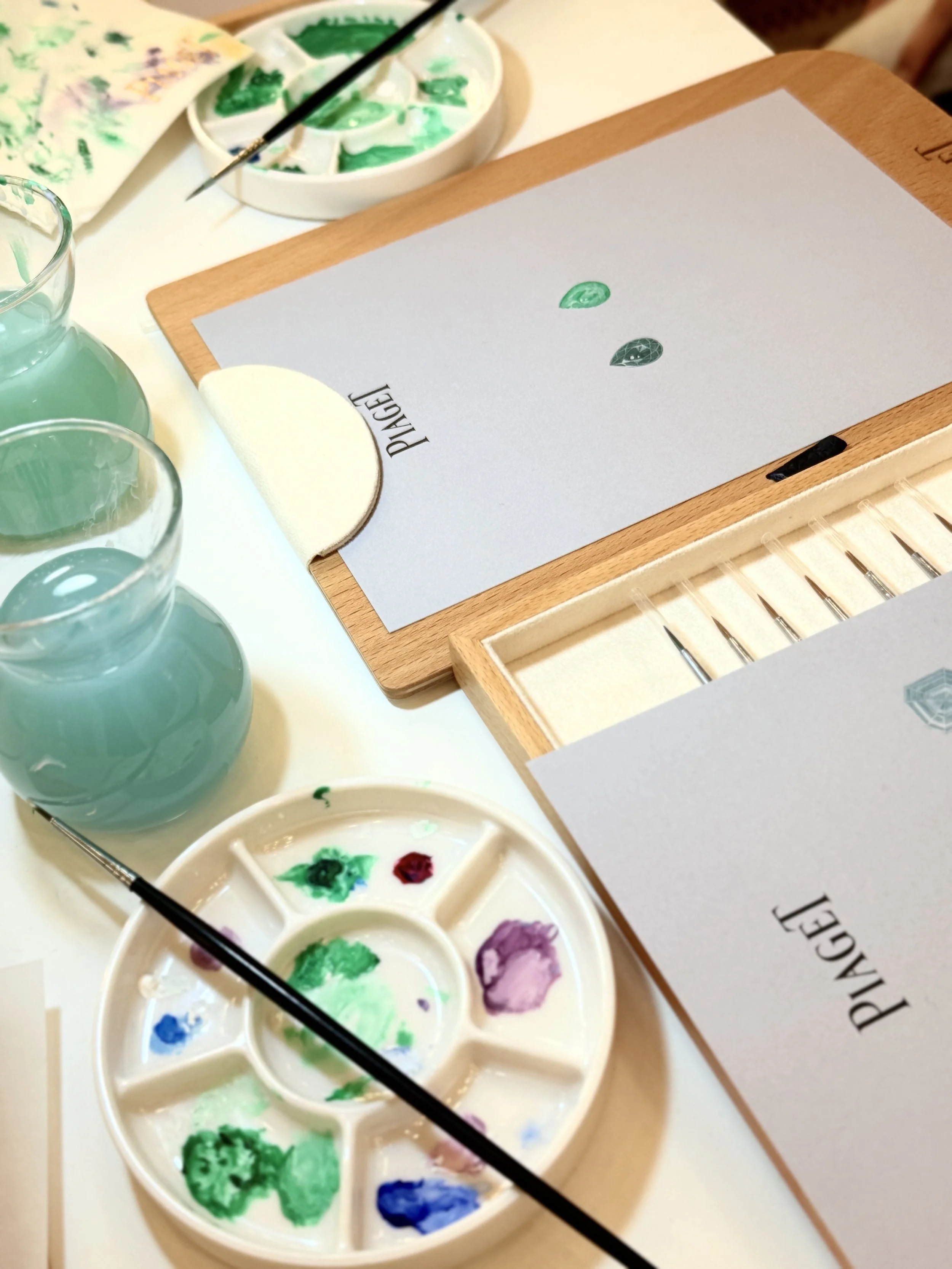 A watercolor palette with green and purple paints, paintbrushes, and a paper with green painted leaves on a desk.