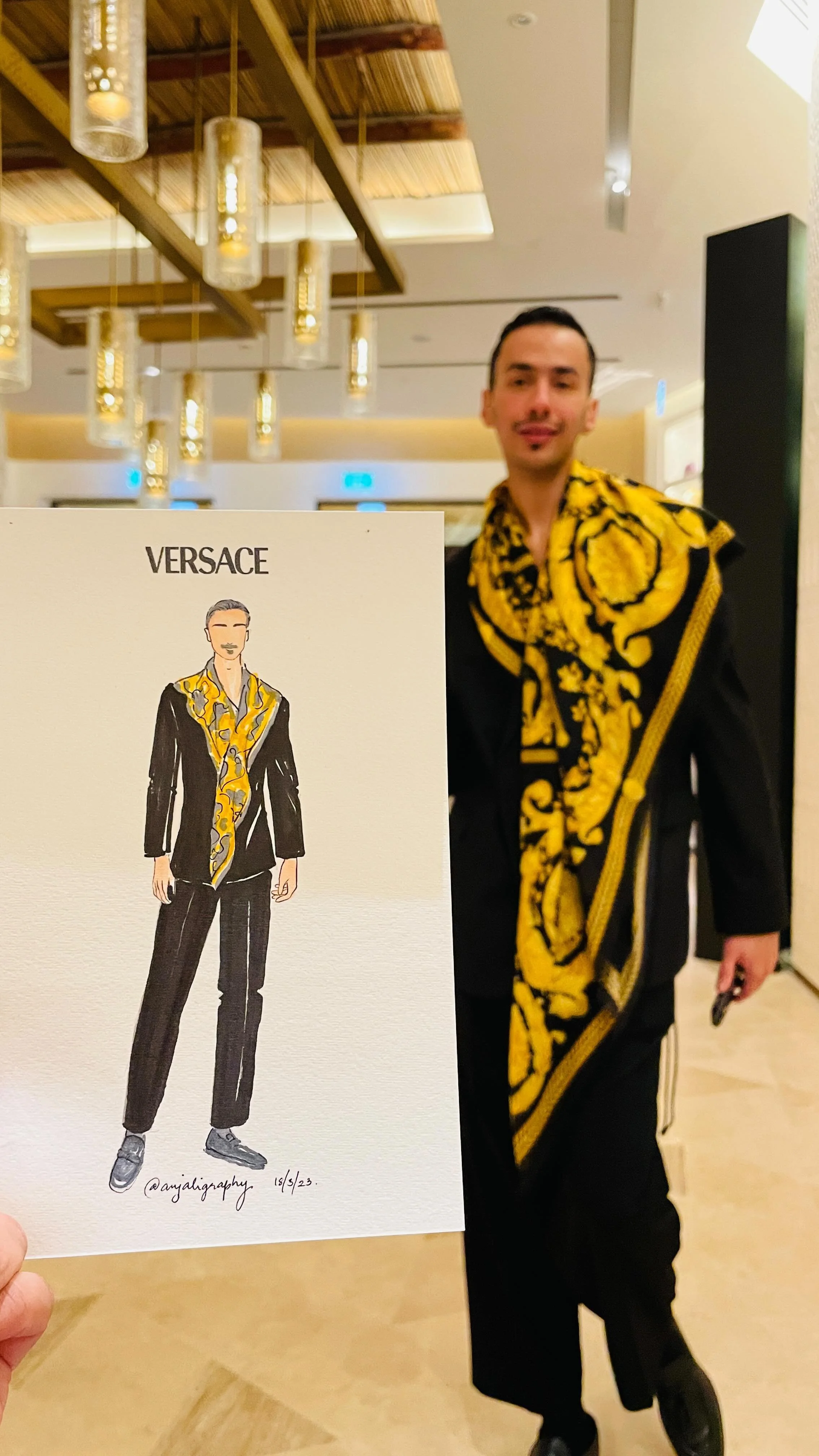 A man wearing a black outfit with a yellow and black patterned scarf stands in a hotel lobby. He is holding a set of keys, and there is a printed fashion illustration of the same outfit in the foreground.