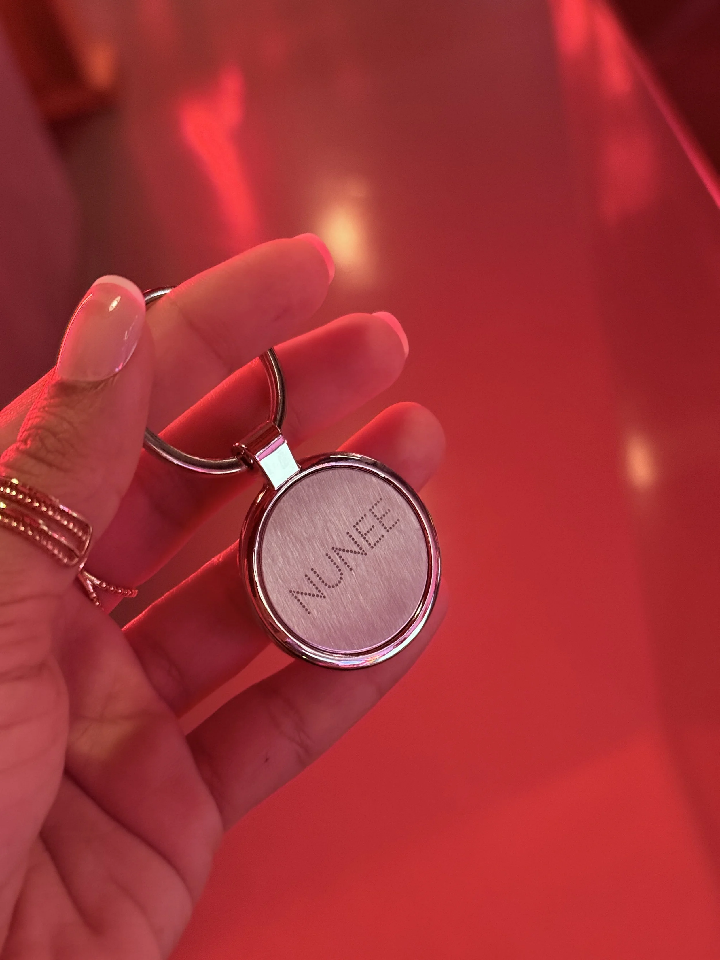 A hand holding a keychain with a metallic round object that has the word 'UNIQUE' engraved on it. The background is red with blurred red and orange lights.