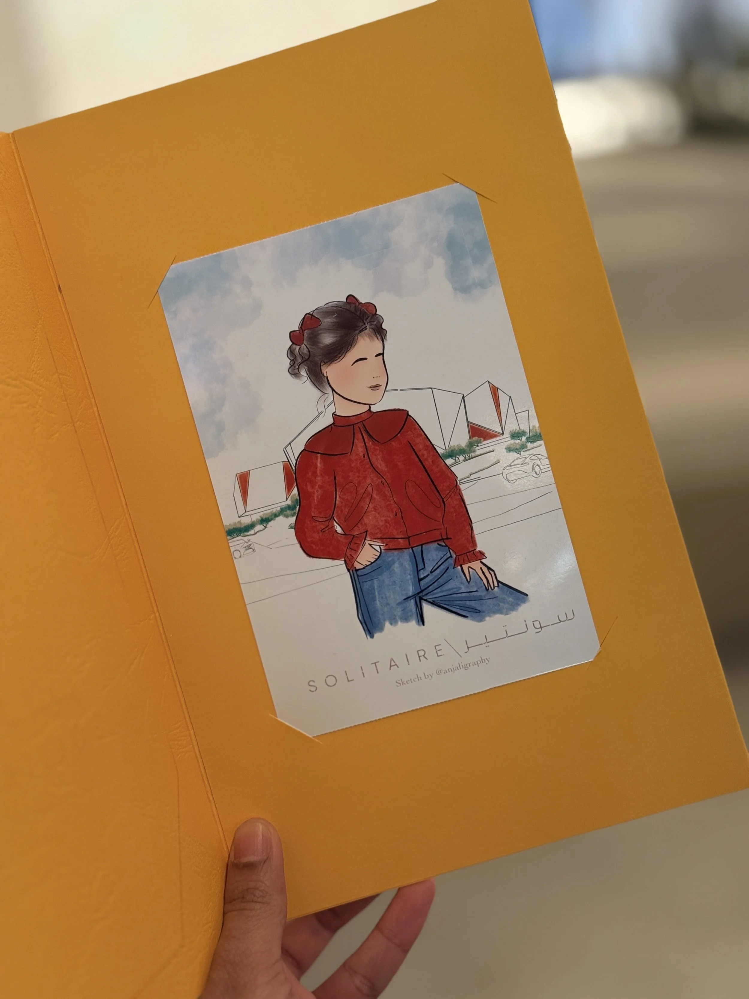 A hand holding a yellow card with a sketch of a woman in a red jacket, with a background of modern buildings and a partly cloudy sky.