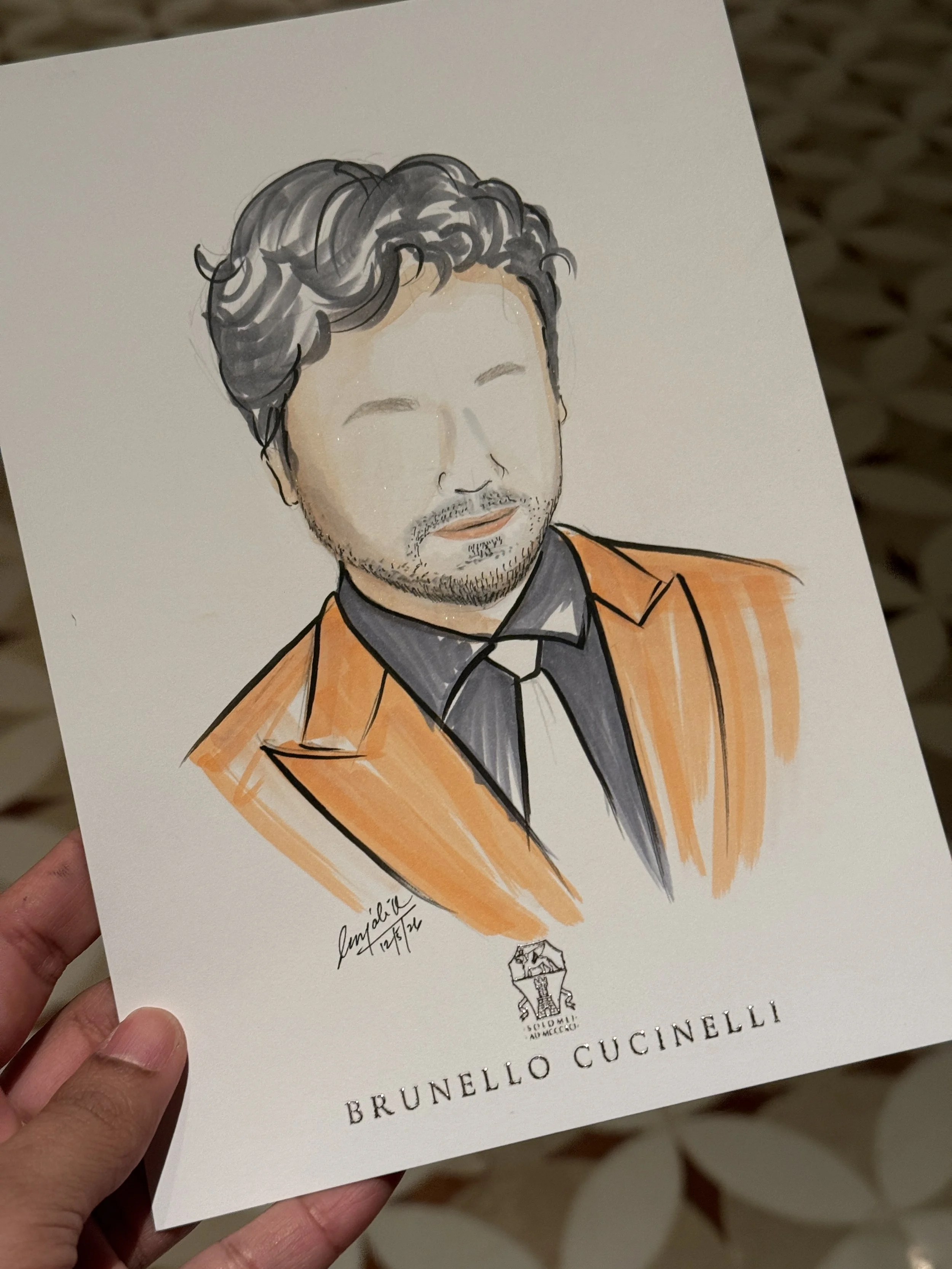 Hand holding a piece of paper with a watercolor portrait of a man with gray, wavy hair and facial hair. The paper features the logo and name of Brunello Cucinelli, a luxury fashion brand.