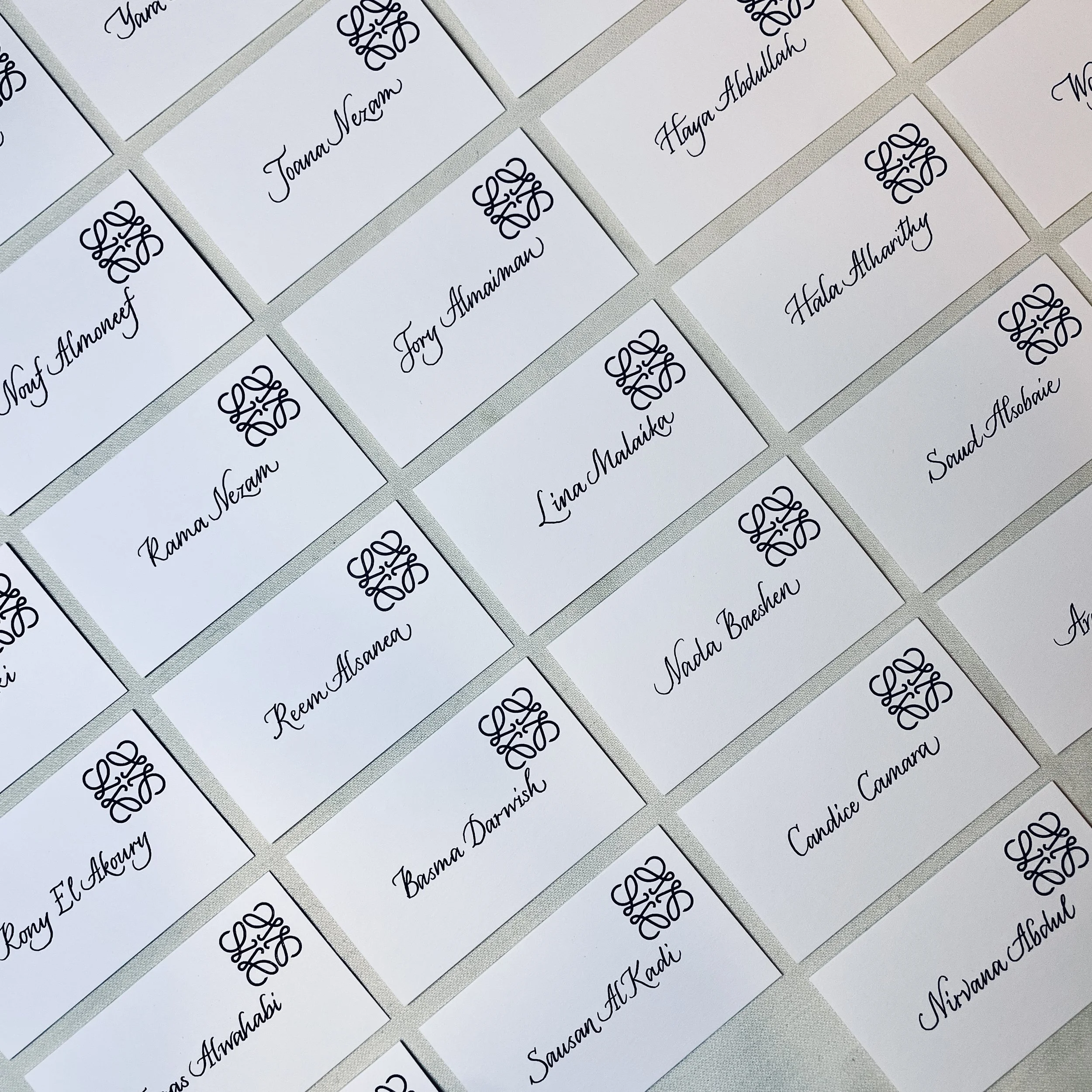 Multiple white name tent cards with names written in black cursive font and a decorative logo above each name, arranged in rows on a light-colored surface.