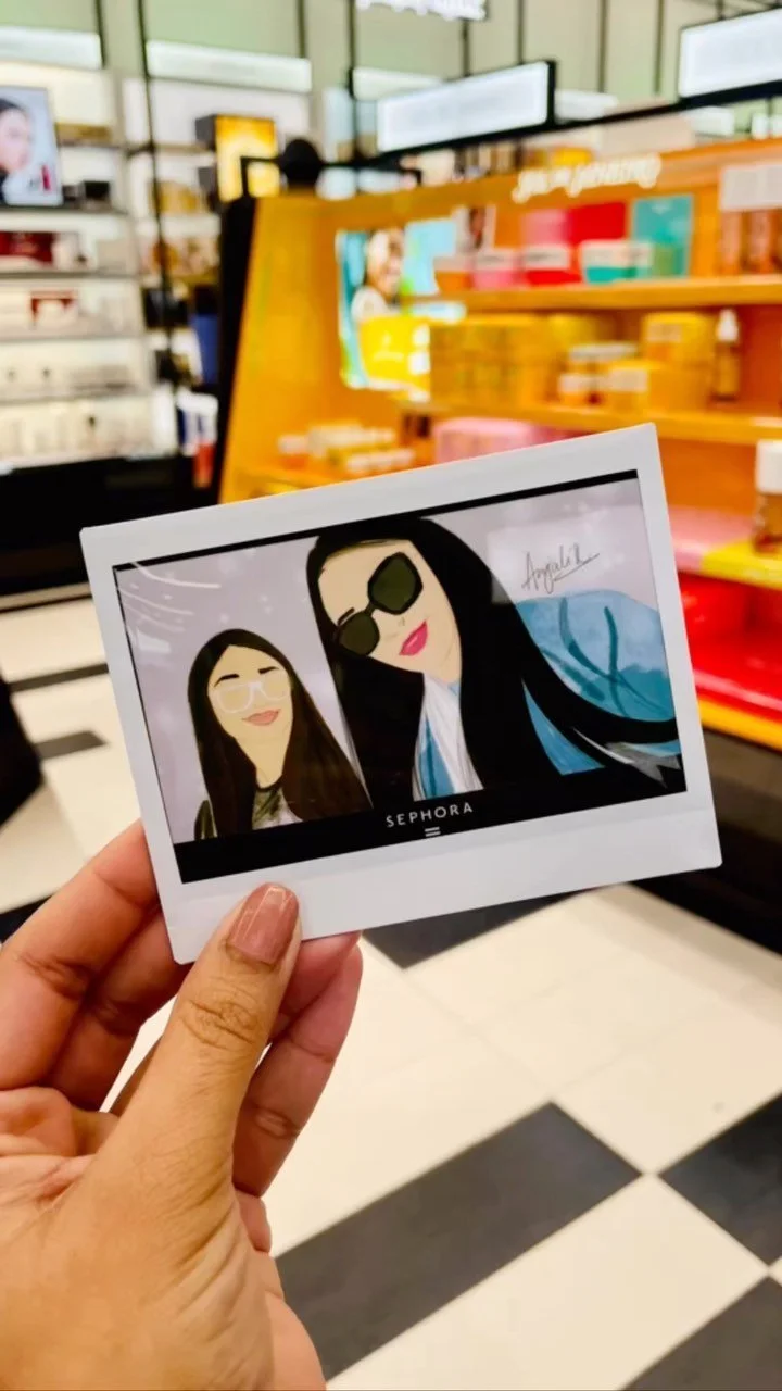 A hand holding a printed cartoon-style photo of two women, one with long dark hair wearing sunglasses and a blue jacket, and the other with long hair and glasses, inside a Sephora store with shelves of beauty products in the background.