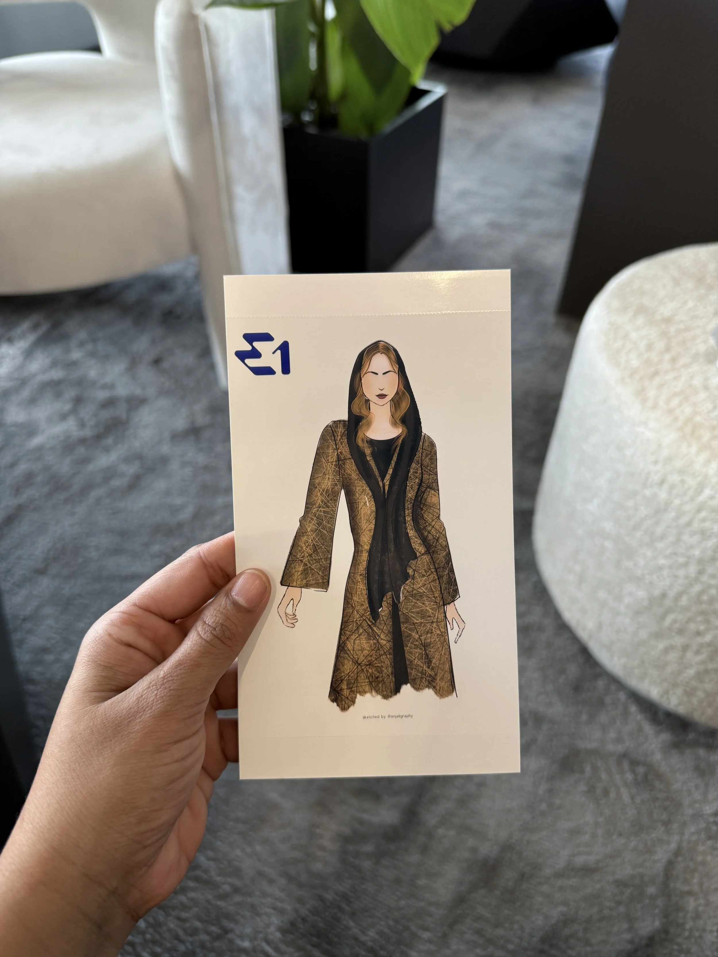 A person is holding a fashion sketch of a woman wearing a long, brown checked coat, black scarf, and black pants, with a black hood. The sketch is on a piece of paper with a small logo in the top left corner and a signature at the bottom.