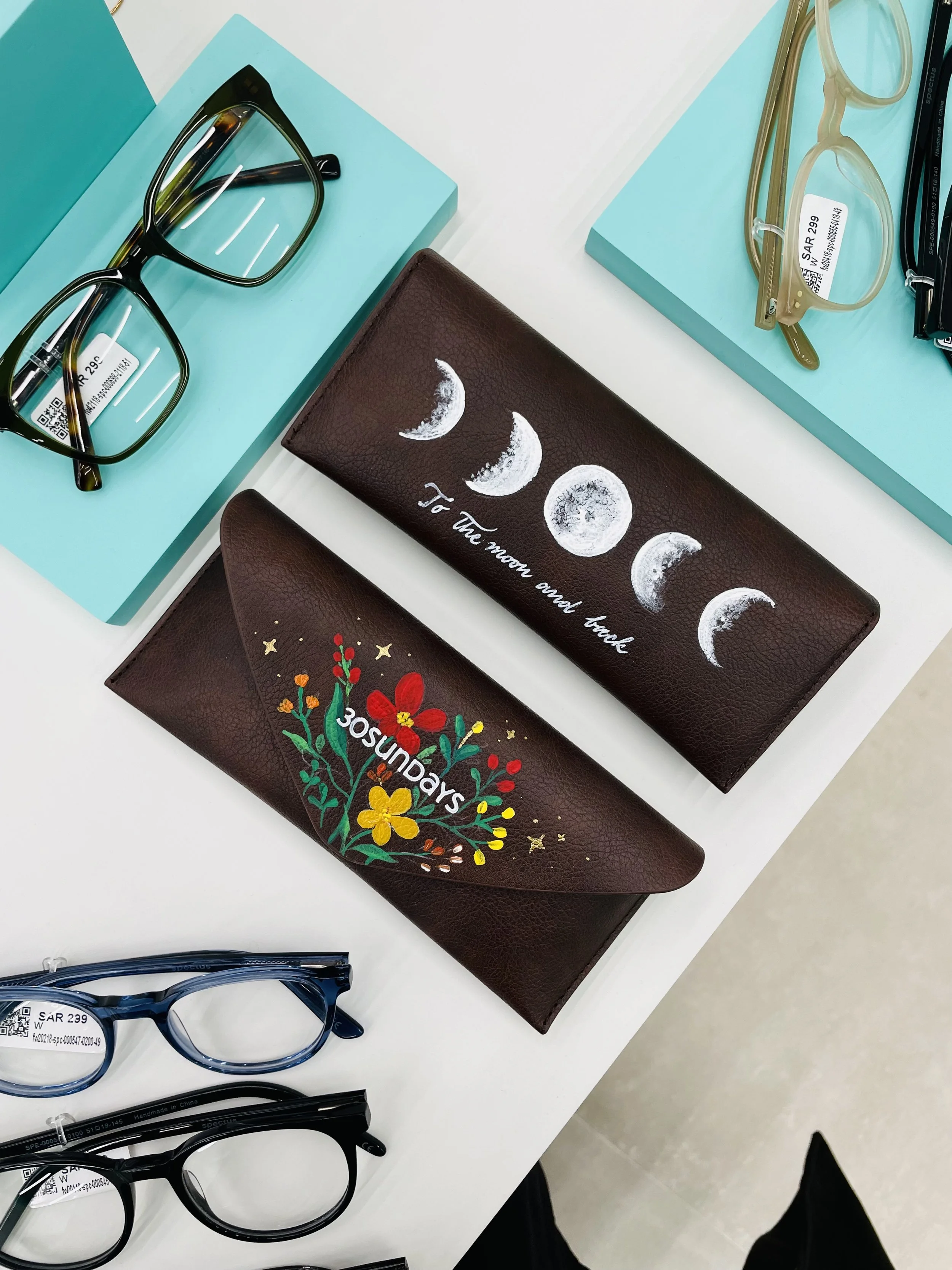 Eyeglasses, one pair with black frame and another with transparent green frame, along with two cloth-covered cases with moon phases and floral designs, displayed on a white surface with light blue boxes.