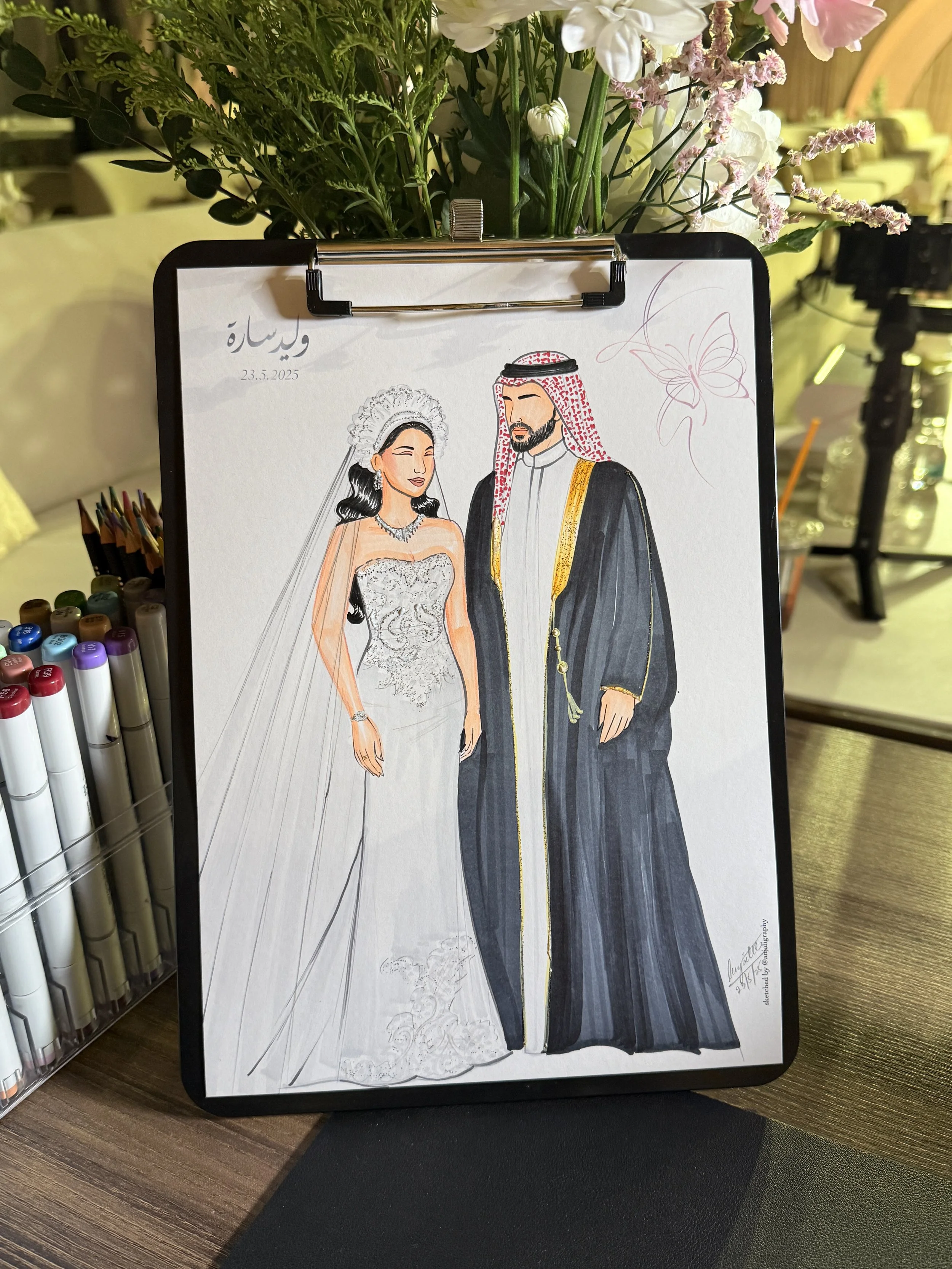 Illustration of a bride and groom in traditional Middle Eastern attire, with the bride in a white wedding dress and the groom in a black robe with gold accents, standing close together on a wedding illustration board surrounded by flowers.