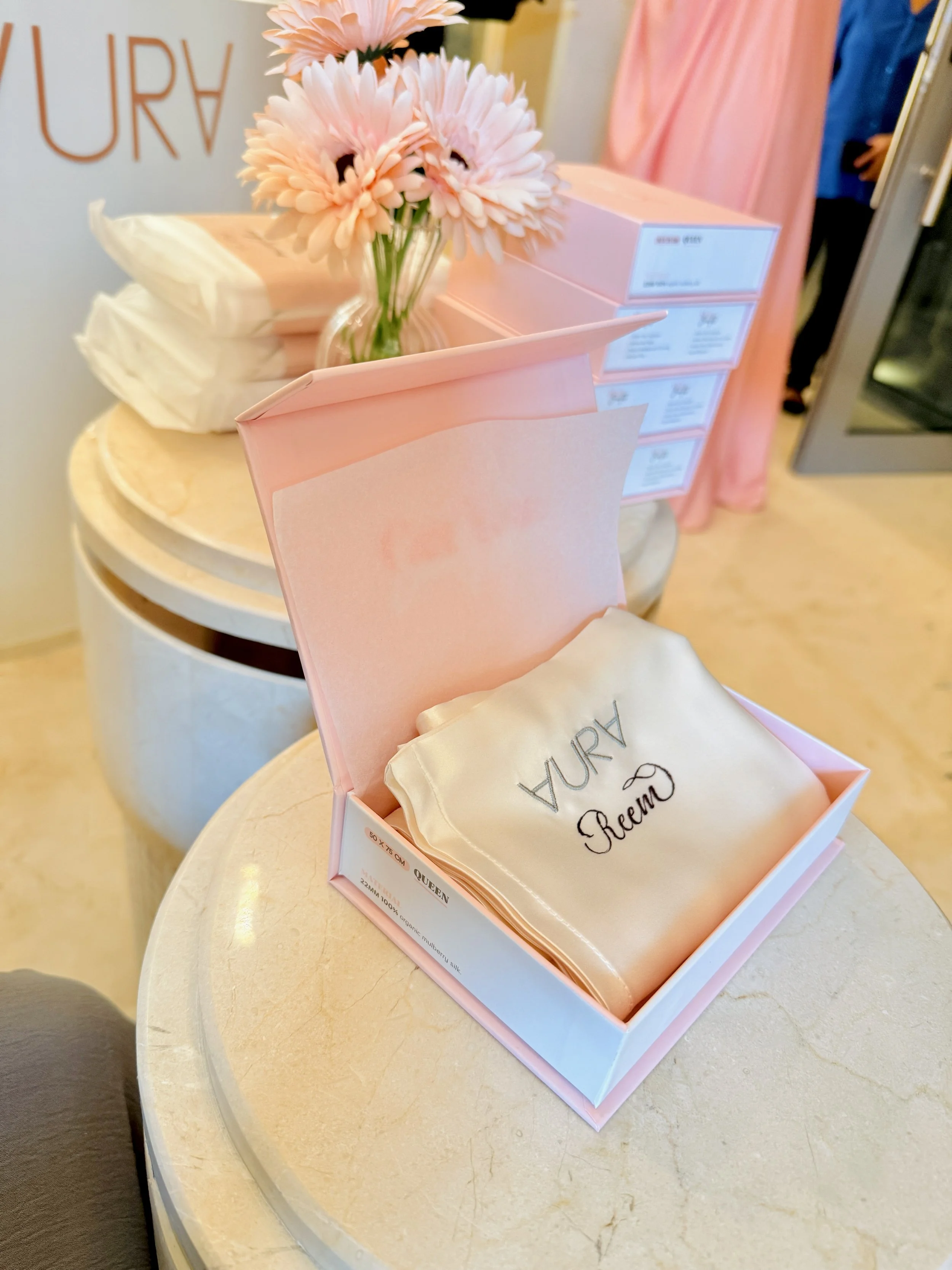 Open pink gift box with a beige cloth inside and the word 'Dream' embroidered on it, placed on a round marble table. In the background, there are pink flowers in a clear glass vase and several stacked pink boxes.