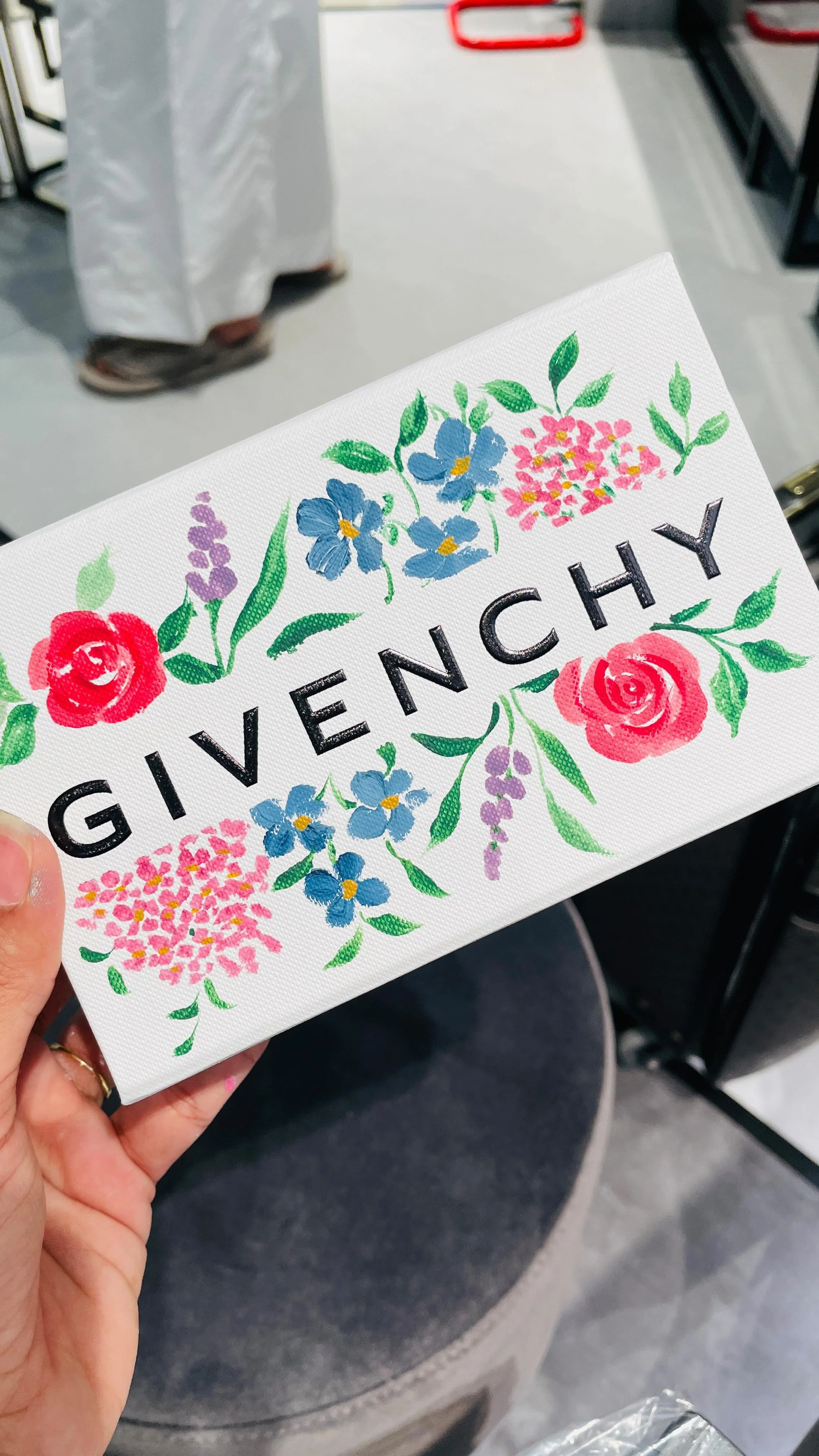 Close-up of a white purse with colorful floral designs and the word 'GIVENCHY' printed on it. The background shows a person's lap and a store environment.