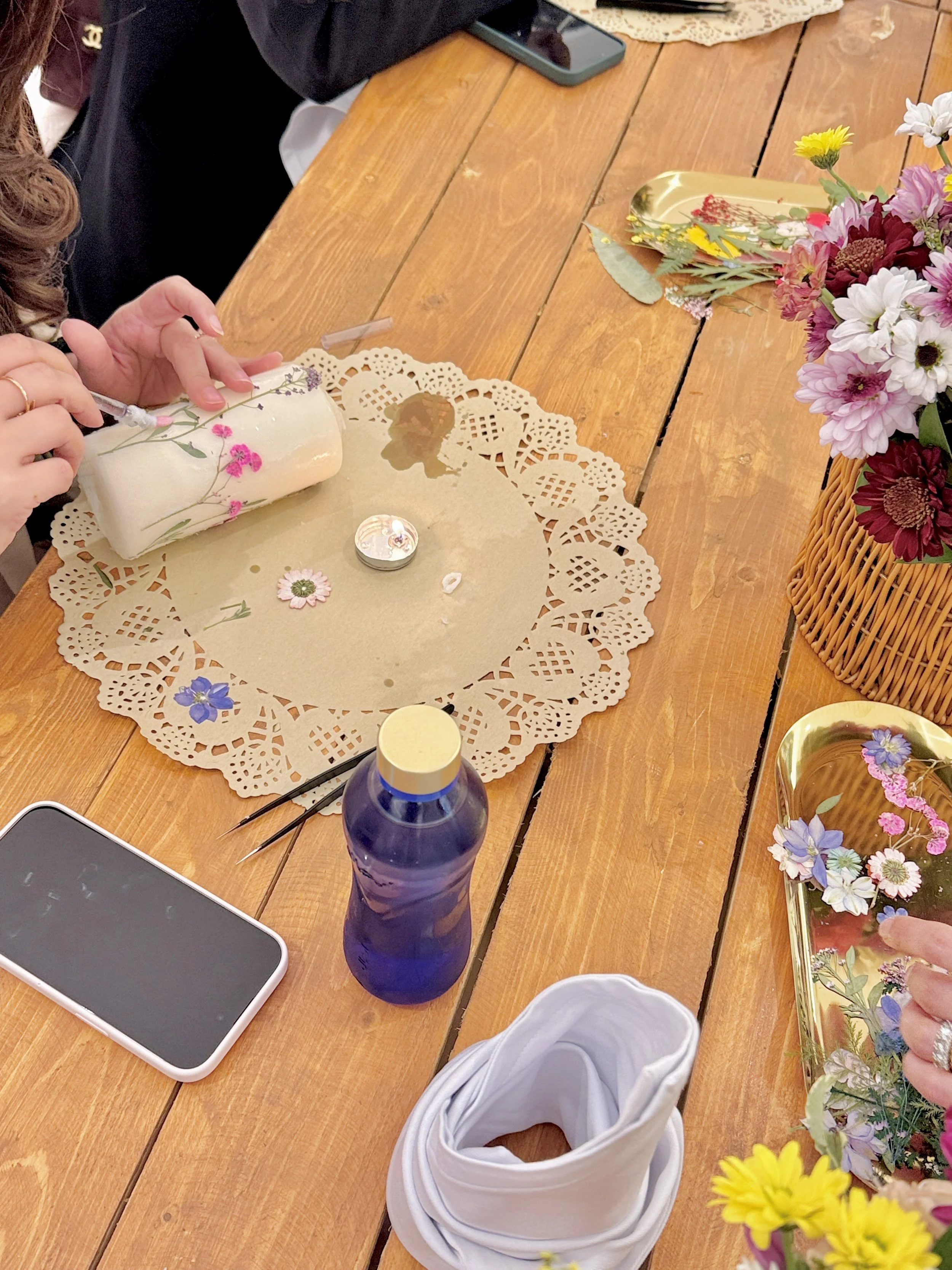 A person decorating a white candle with small purple and pink flowers on a wooden table with flowers, a phone, a candle, and a bottle nearby.