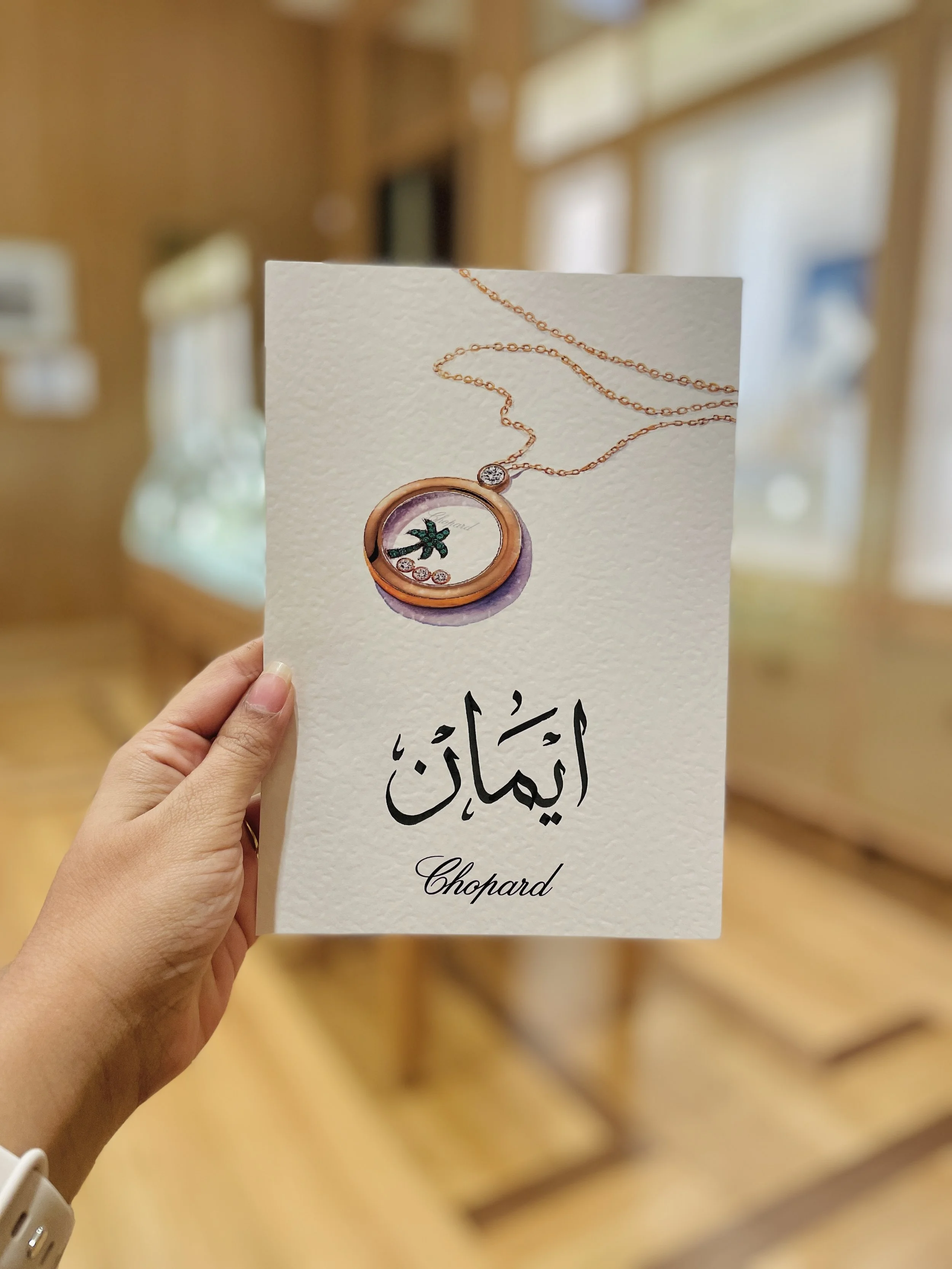 A person holding a white card with a picture of a rose gold necklace featuring a circular pendant with a flower design and small diamonds, with Arabic calligraphy and the word 'Chopard' underneath.