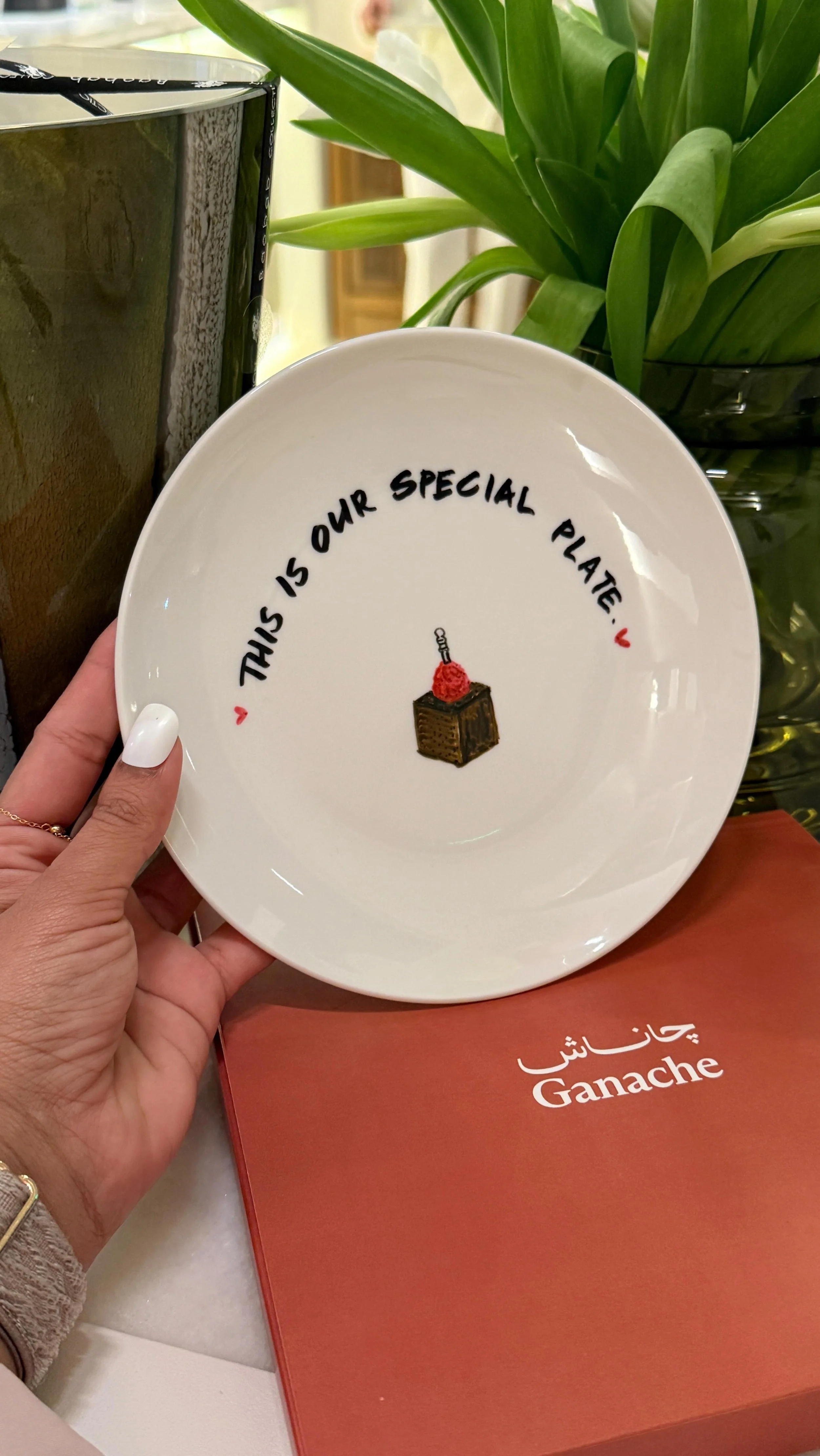 A white plate with black text reading 'This is our special plate!' and a small illustration of a cake with a cherry and a candle. A person's hand holding the plate is visible. There is a potted plant with green leaves and a box of Ganache chocolates 