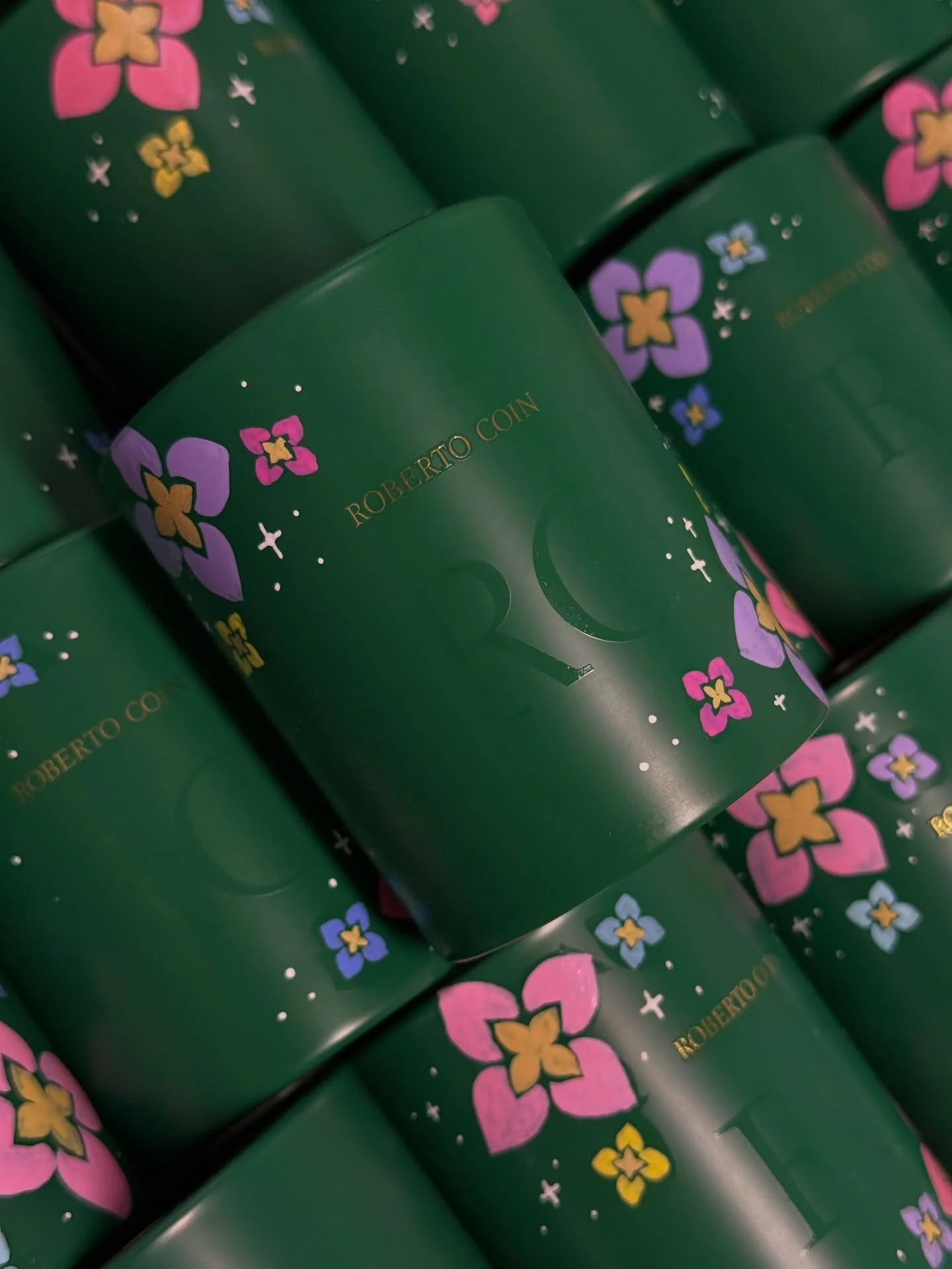 Close-up of green cylindrical containers with the words 'ROBERTO COIN' written in gold, decorated with pink, purple, and yellow floral designs and small white stars.