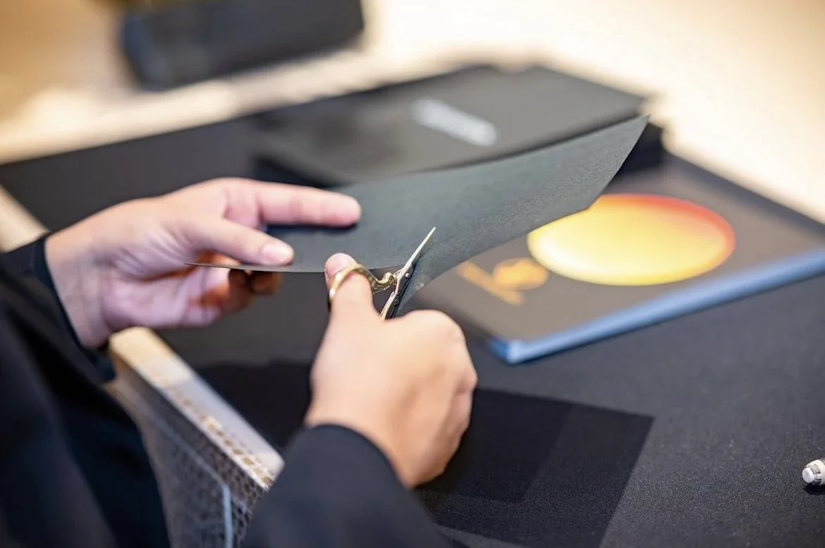Close-up of hands cutting black paper with scissors over a table with a black surface, a colorful book, and a crayon.
