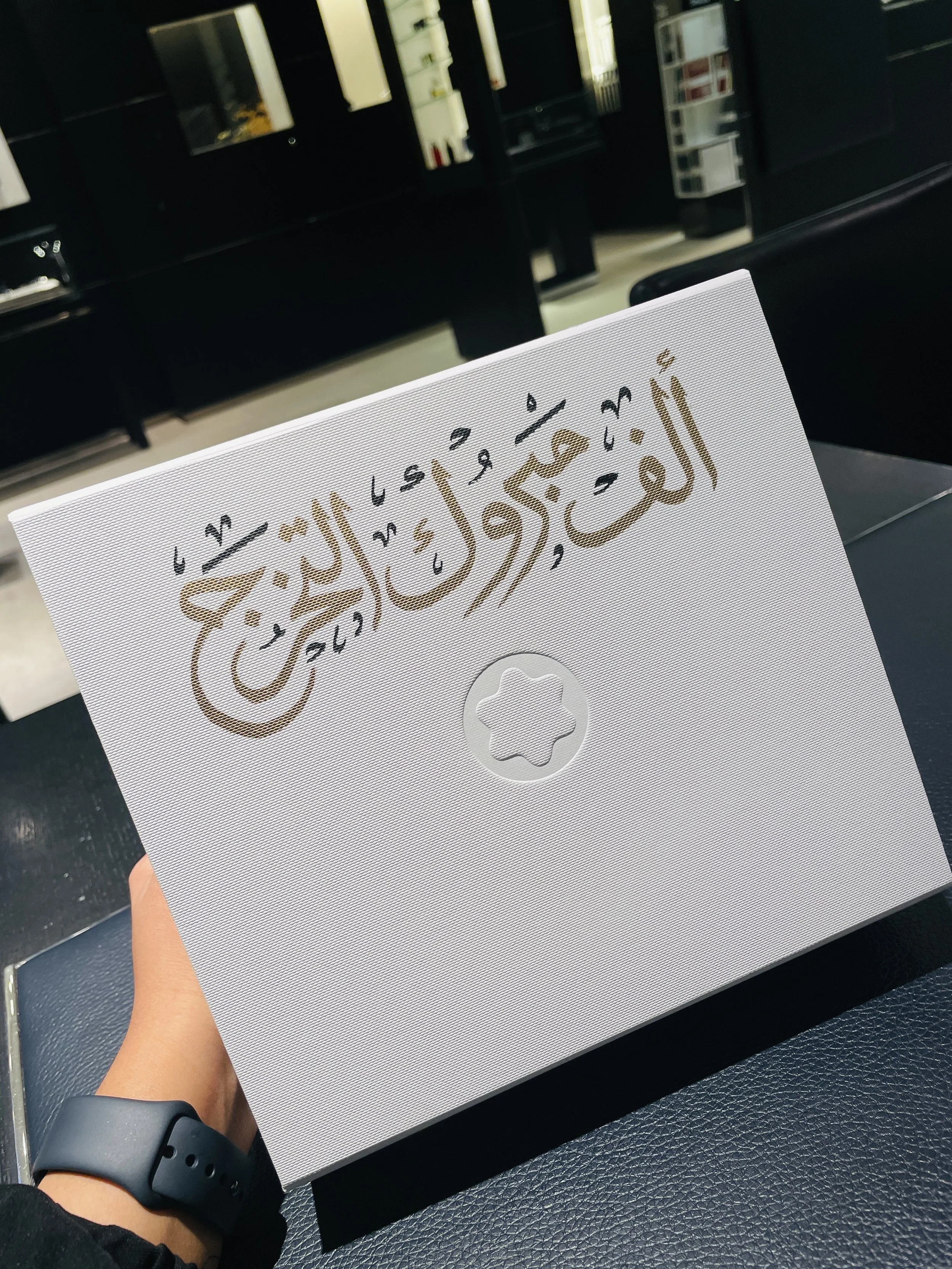 A person holding a white card with Arabic calligraphy and a circular hologram, with a dark shelf and pharmacy supplies in the background.