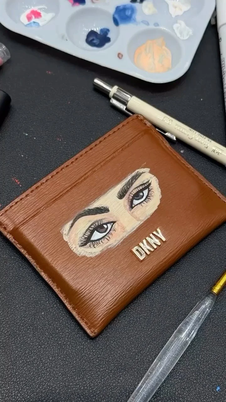 Close-up of a brown leather wallet with a painted portrait of expressive blue eyes and black eyebrows, and gold initials "DKNY." Nearby are art supplies, including paint palette, white paint pen, and a precision screwdriver.