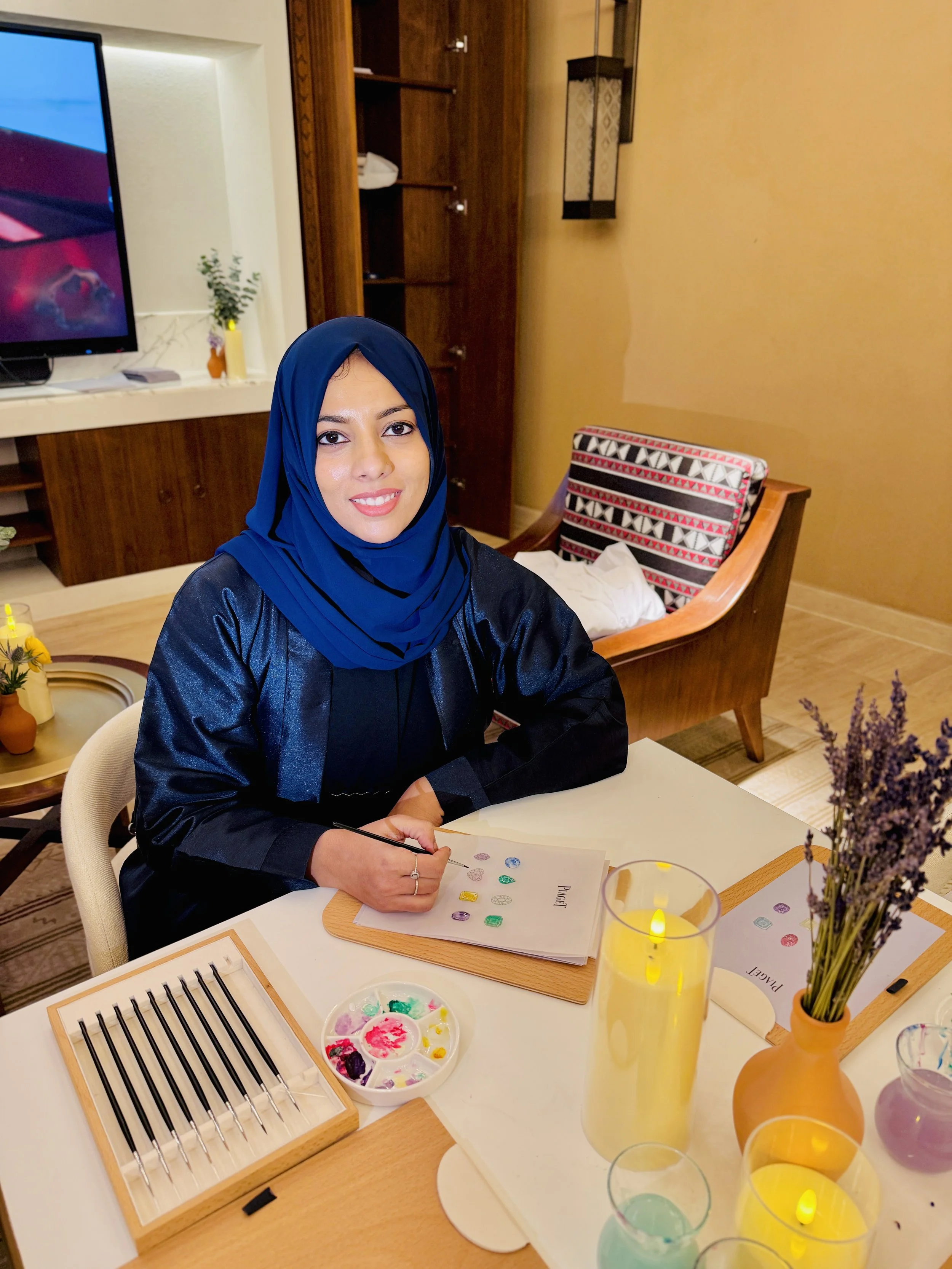 A woman in a blue hijab and black top sitting at a table with painting supplies, including a paint palette, brushes, and glass vials with colorful liquids. She is smiling and holding a paintbrush. The background shows a brown wooden cabinet, a TV, an