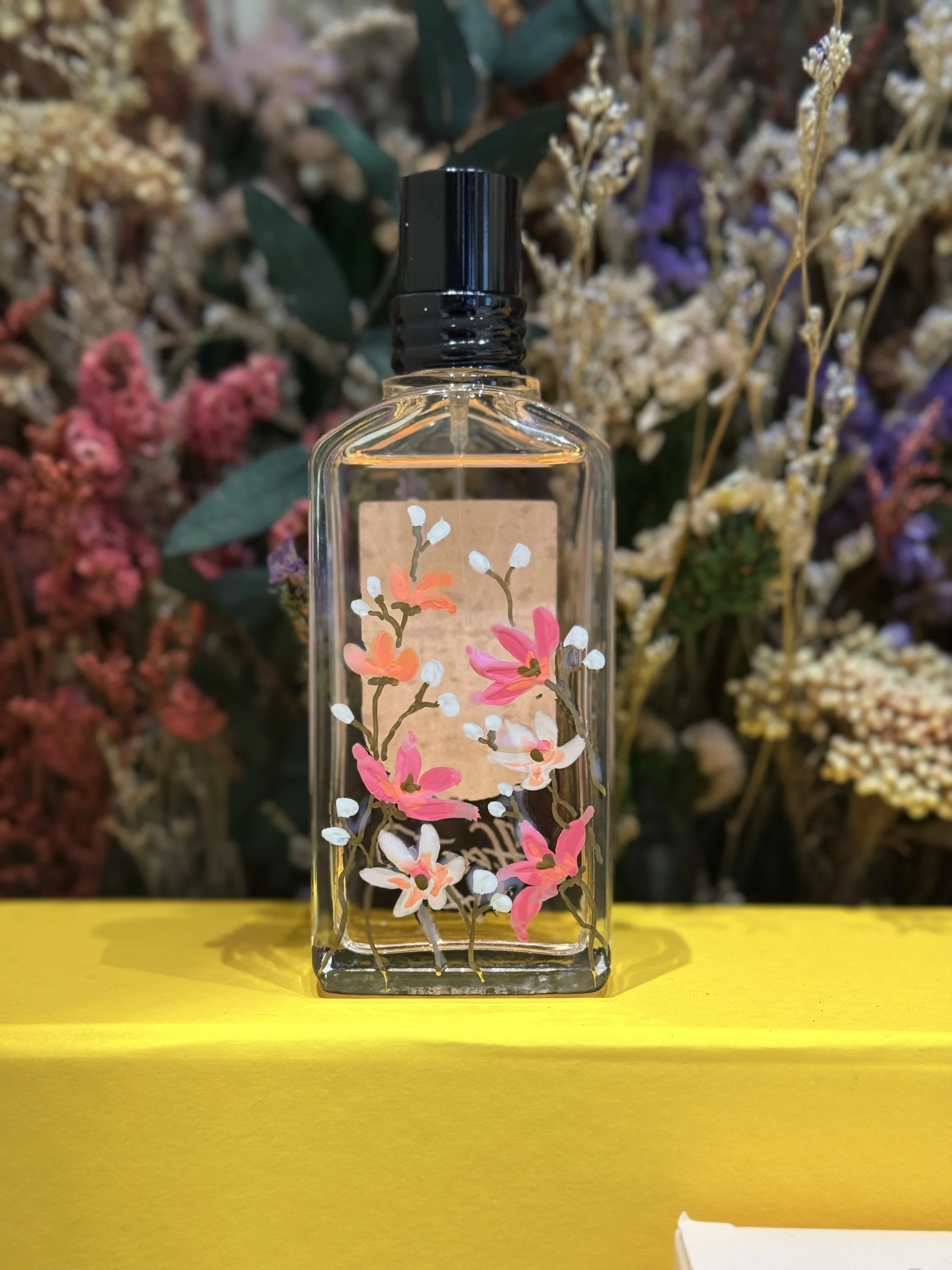 A clear glass spray bottle with a black cap, decorated with painted pink and white flowers, is placed on a yellow surface with a background of colorful flowers.