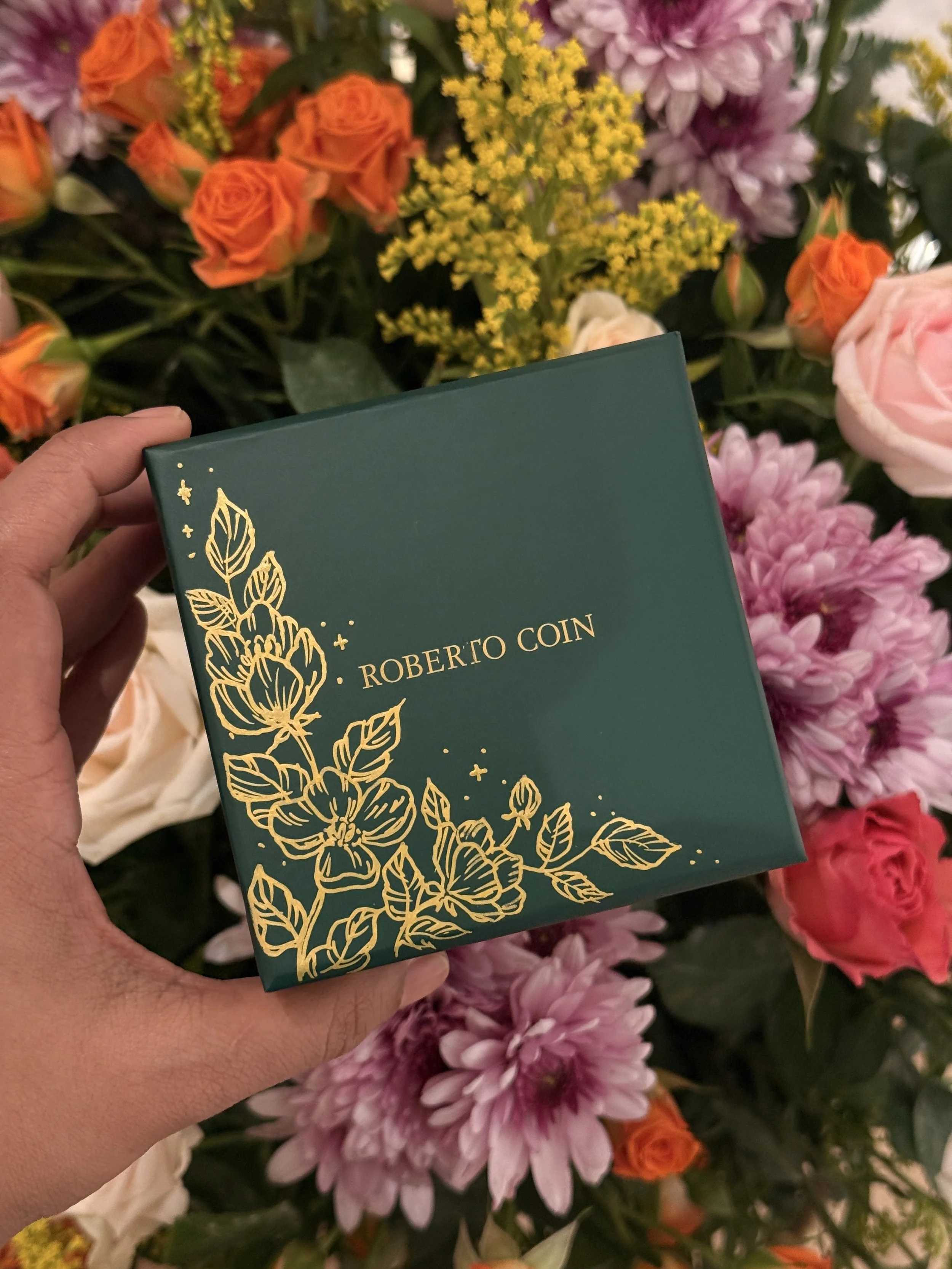 A hand holding a green box with gold floral design and text, in front of a colorful display of pink, orange, yellow, and purple flowers.
