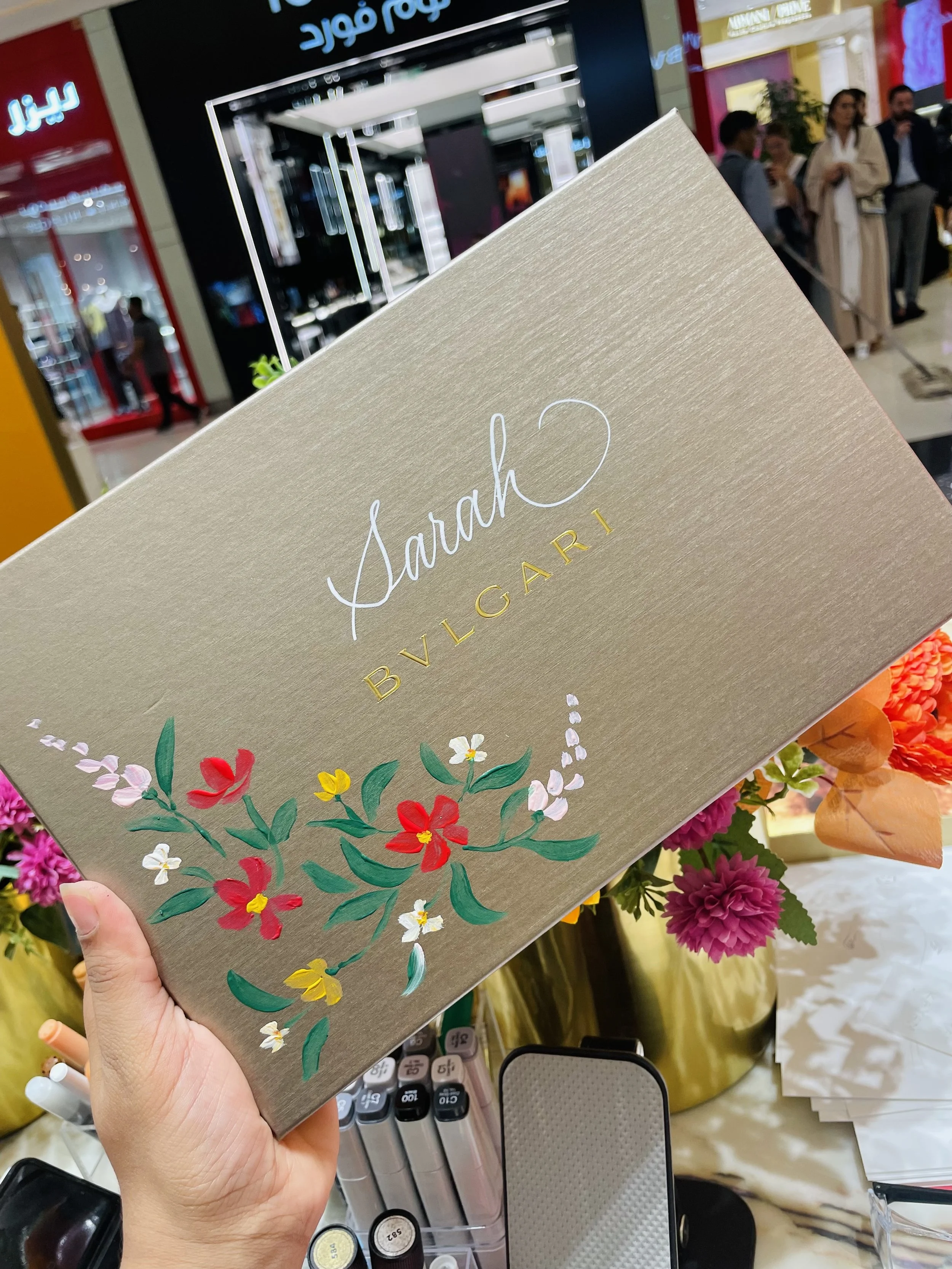 A hand holding a beige book with the title 'Sarah' and the word 'Bulgari' embossed in gold, decorated with colorful floral illustrations, in a store with various products and people shopping in the background.