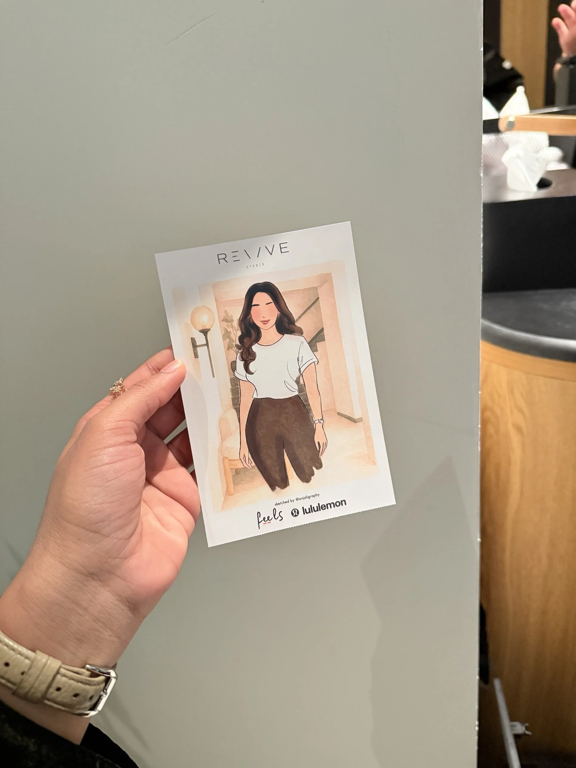 Hand holding a ticket with an illustrated woman in casual clothing, standing in a warmly lit interior with a lamp, plant, and staircase. The ticket has the words 'feels' and 'lululemon' at the bottom.