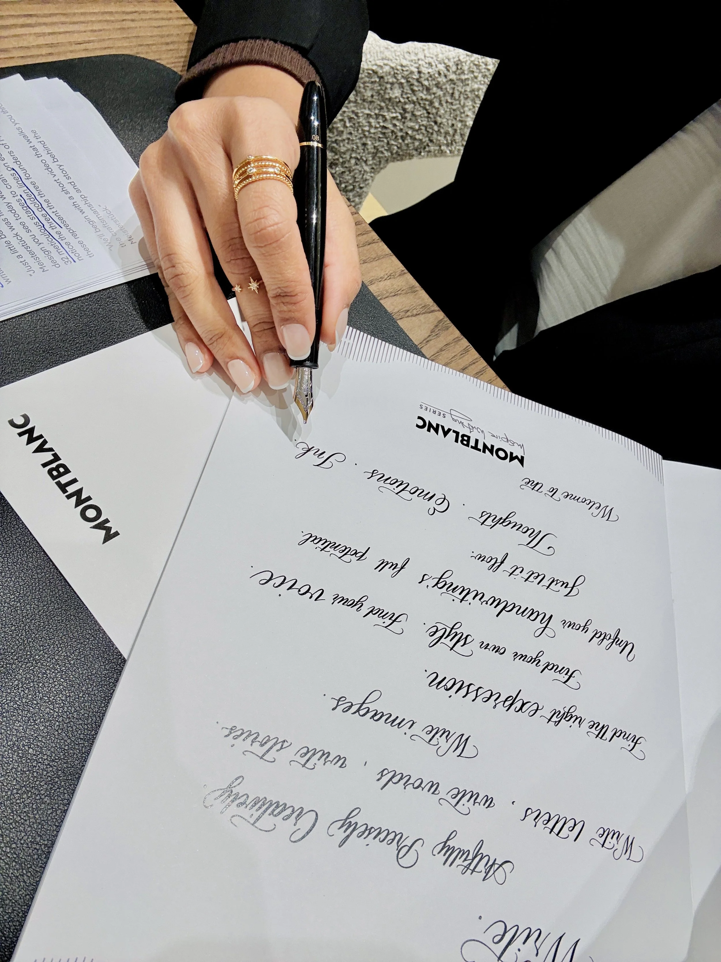 Close-up of a hand holding a fountain pen, signing a document on a black table. The hand is wearing multiple gold rings and a star-shaped ring, with neatly manicured nails. A stack of papers is partially visible on the left. The document has elegant 