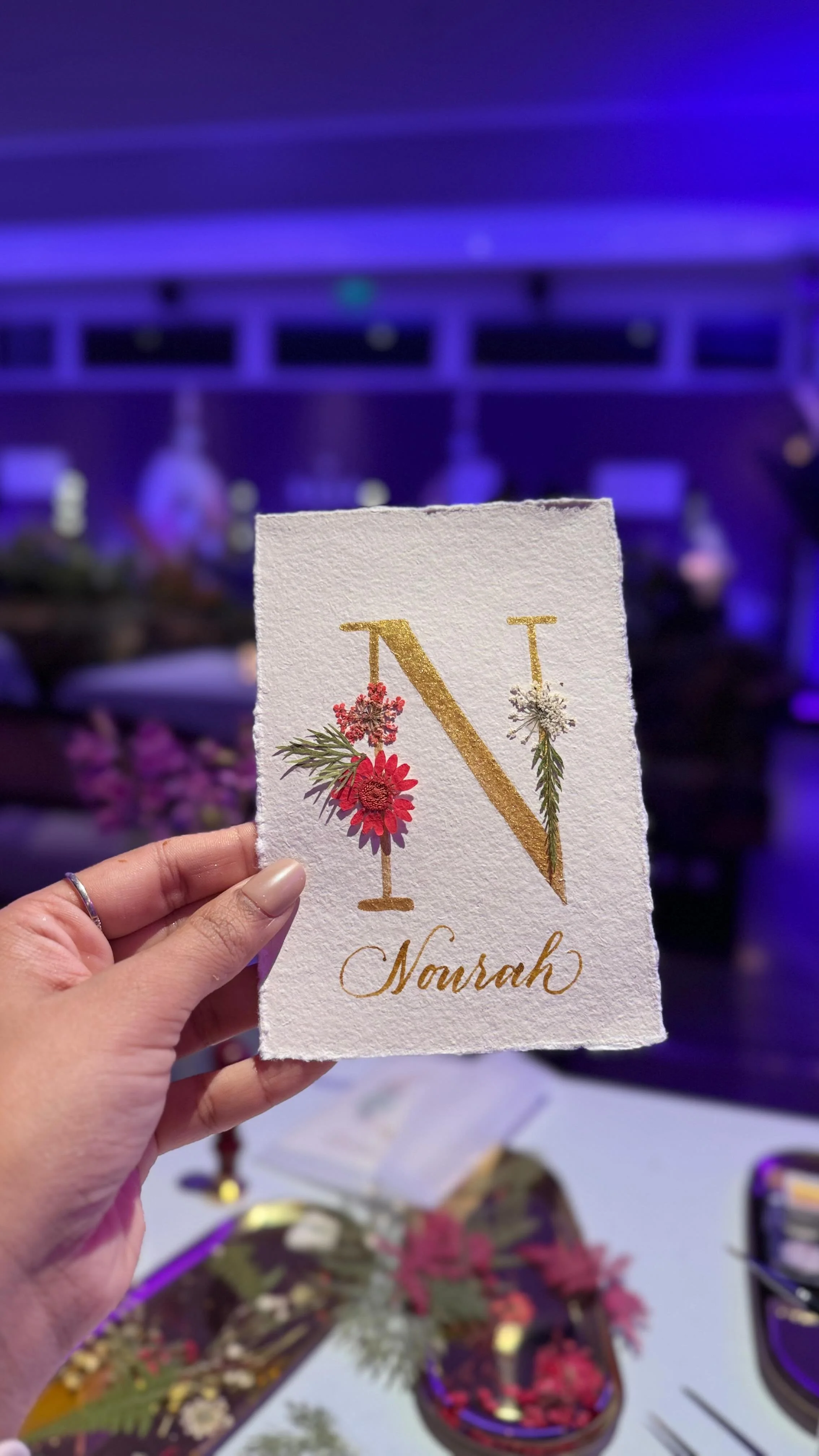 A person holding a handmade card with the letter N decorated with flowers, with a blurred background of a table with craft supplies and a purple-lit setting.