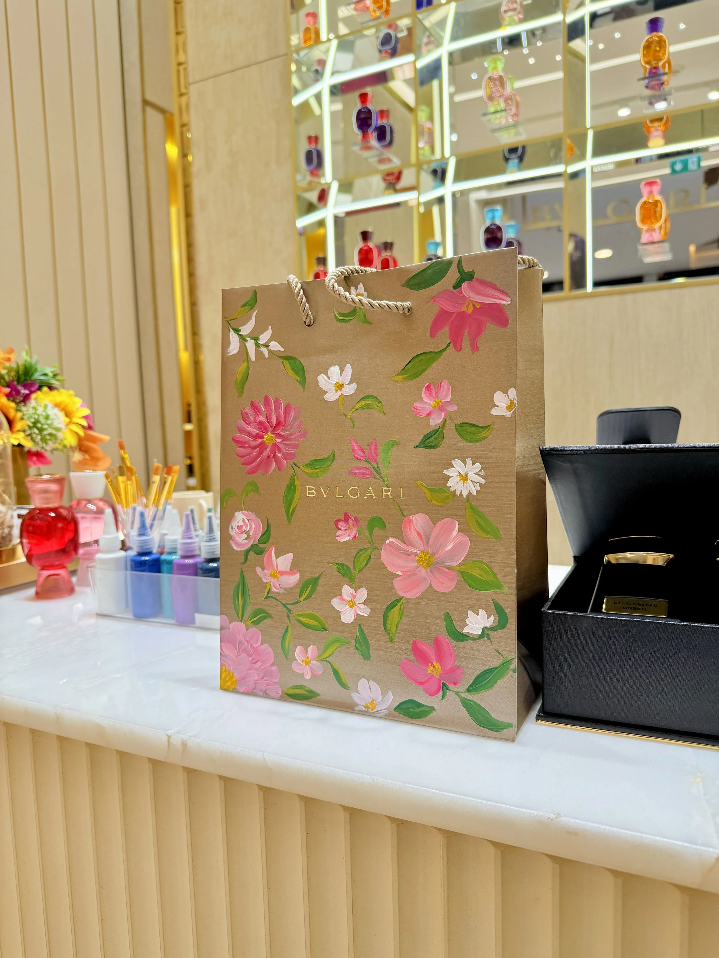 Bvlgari shopping bag with pink and white floral design on a white display table, surrounded by small colorful bottles and framed by a wall with glass shelves filled with perfume bottles.