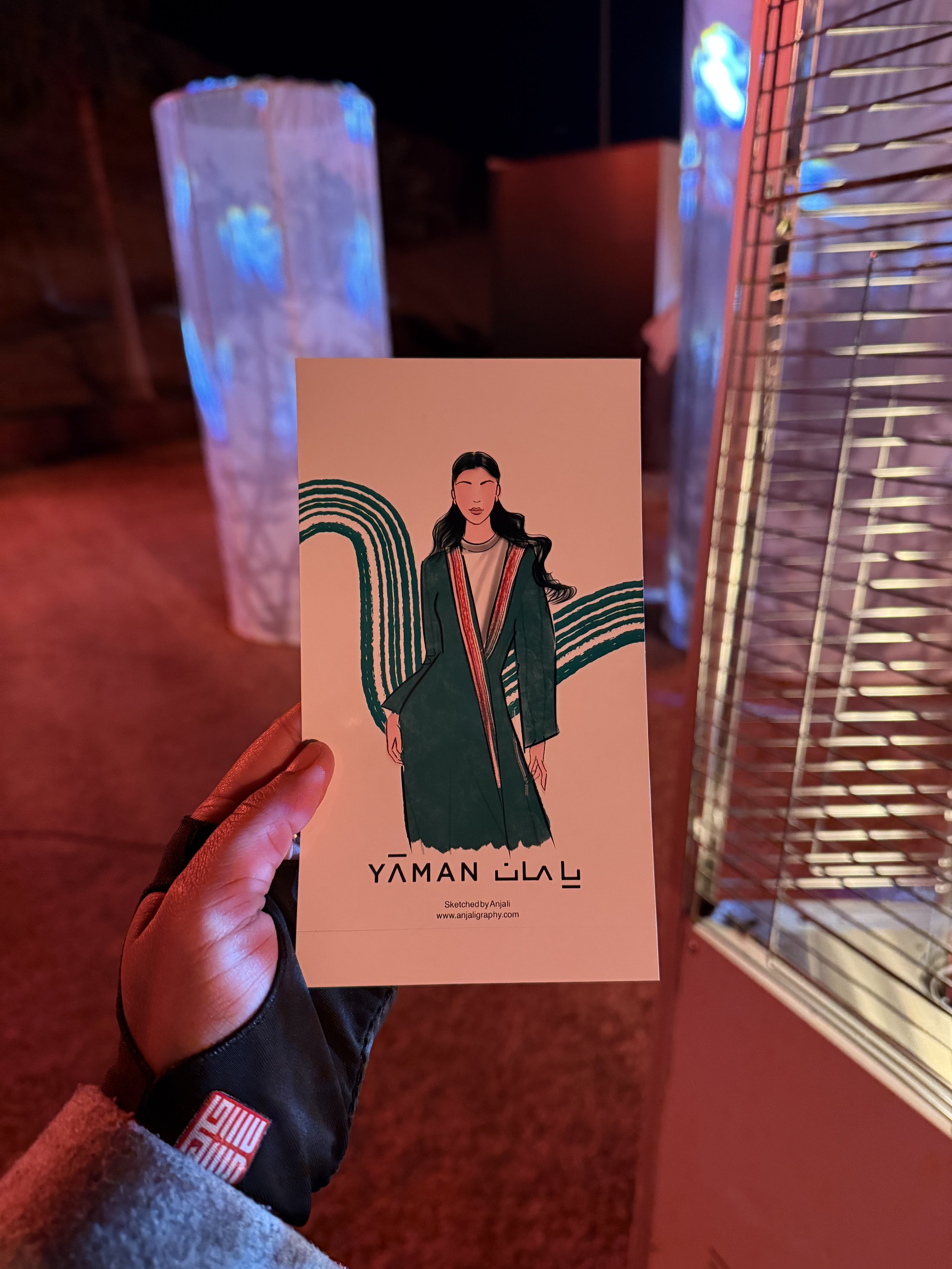 A hand holding a Yamin Gal poster featuring a woman in traditional attire with a pink background, in an indoor space with illuminated light fixtures and red flooring.