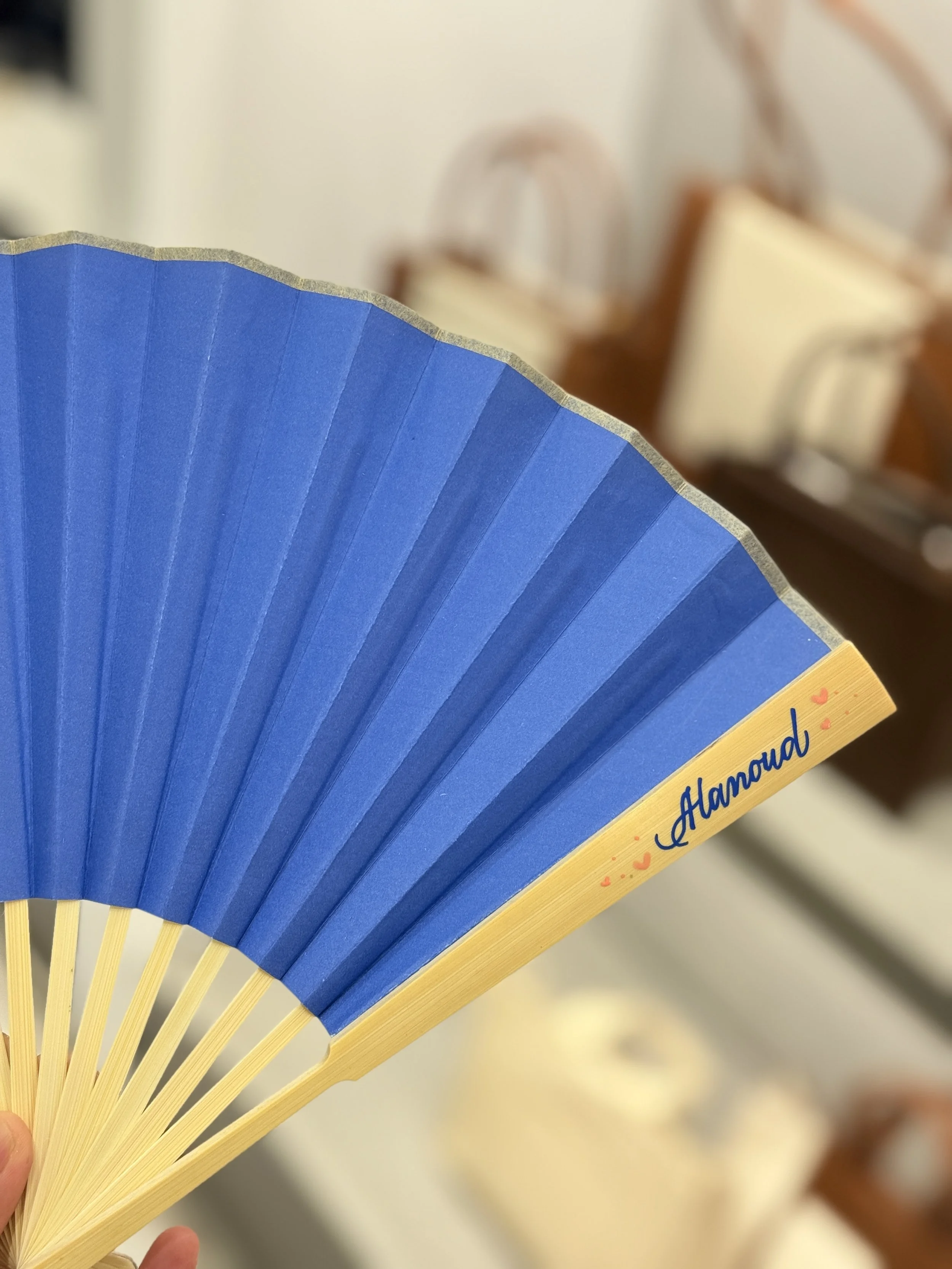 Close-up of a blue hand fan with yellow ribs, with the word 'Glamour' and small heart designs on the yellow edge.
