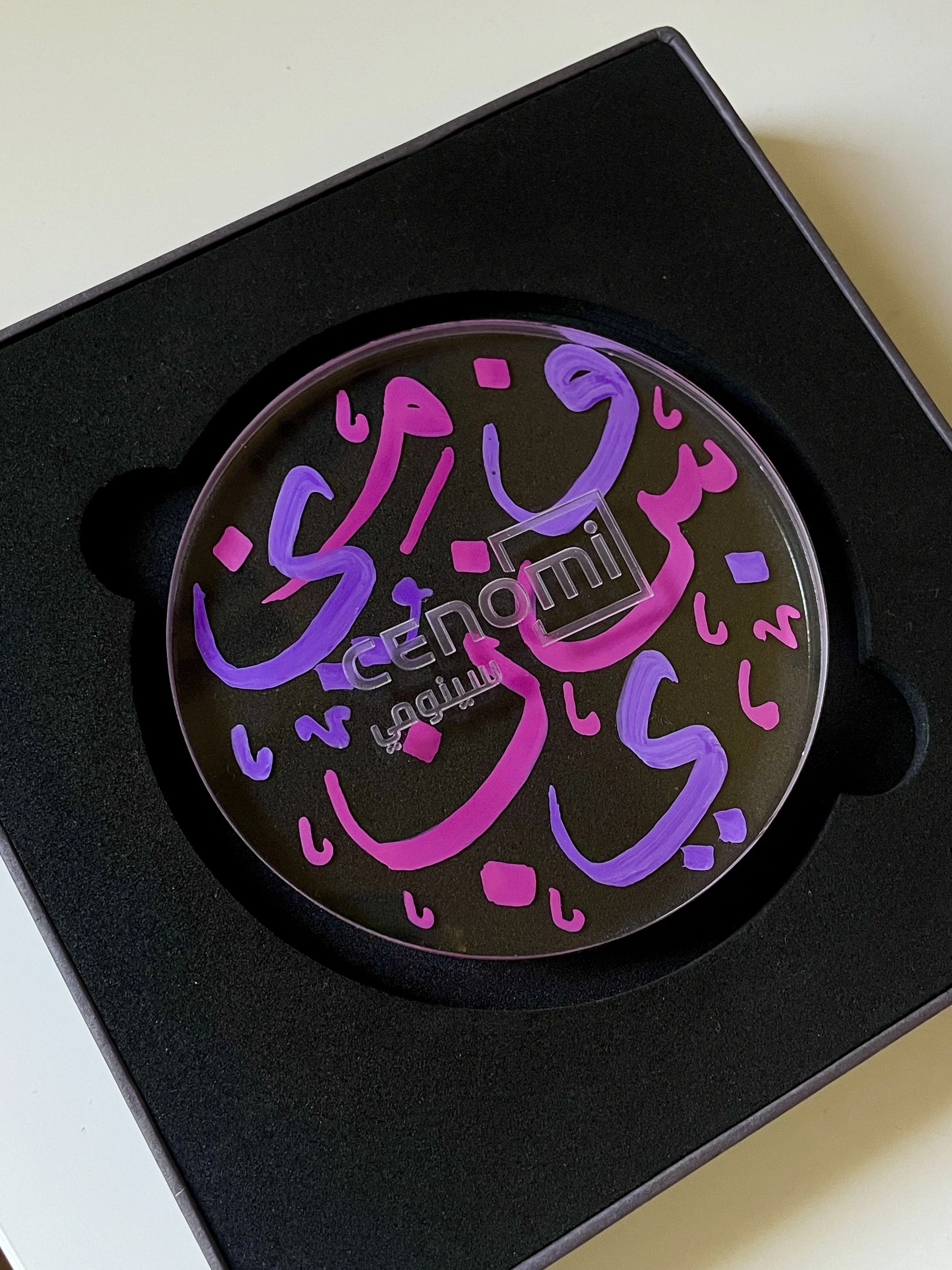 Close-up of a round black surface with colorful pink and purple scribbles and a small transparent square with the text 'emi' on it, placed on a box or frame.