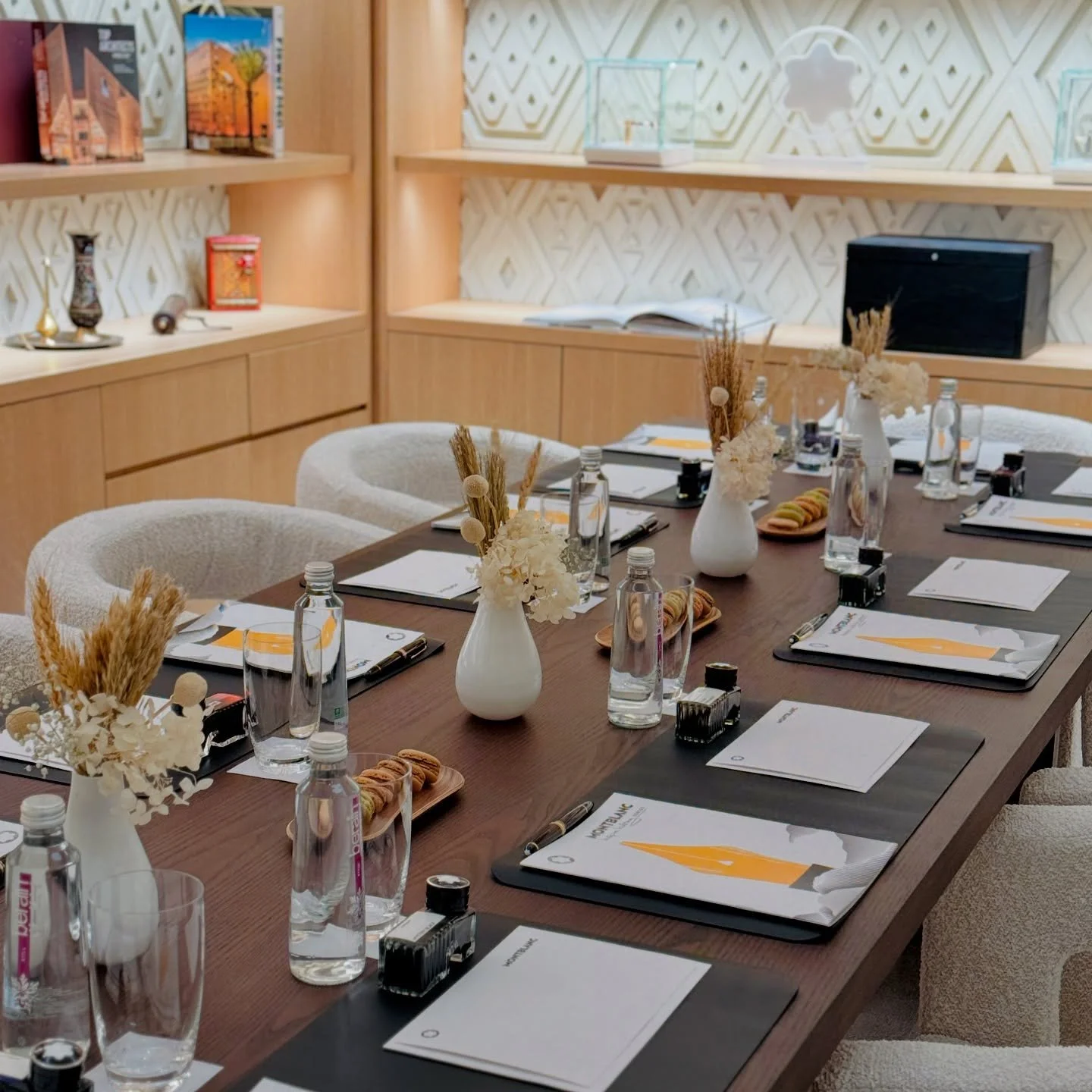 A conference or meeting room with a long wooden table set with notebooks, pens, water bottles, and small flower arrangements in white vases. The room has beige, textured chairs and wooden shelves with books and decorative objects.