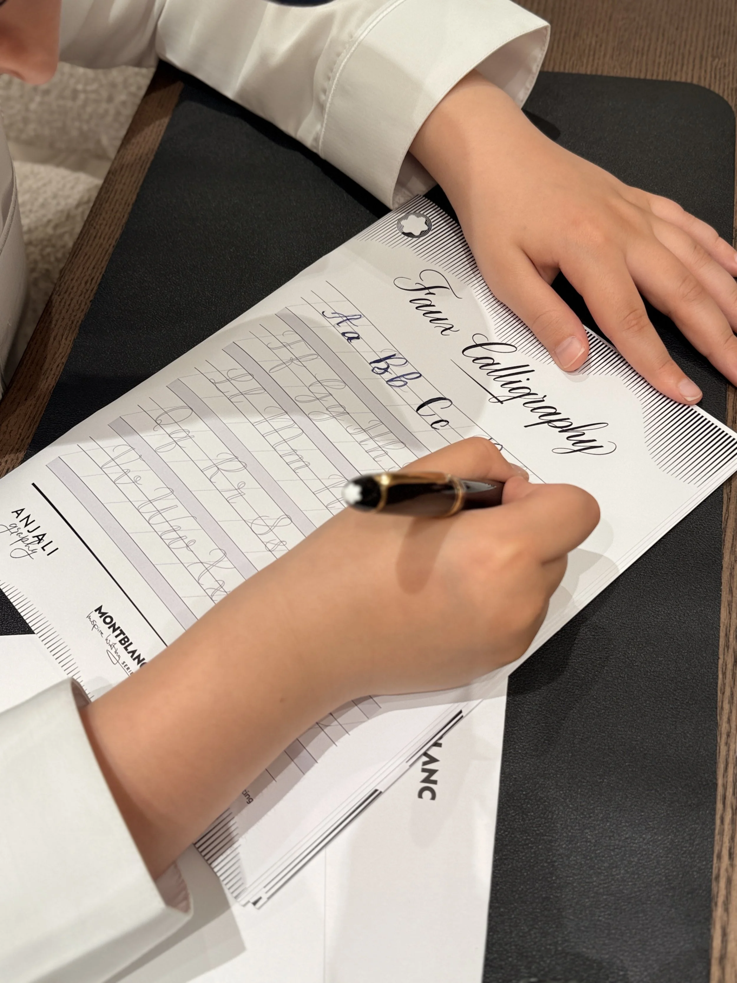 A person's hand holding a pen, practicing calligraphy on a sheet of paper that says 'Fawn Labigrow' and 'Have Bb Ce' with other handwritten cursive notes. The paper is on a black surface, and the person is wearing a cream-colored shirt.