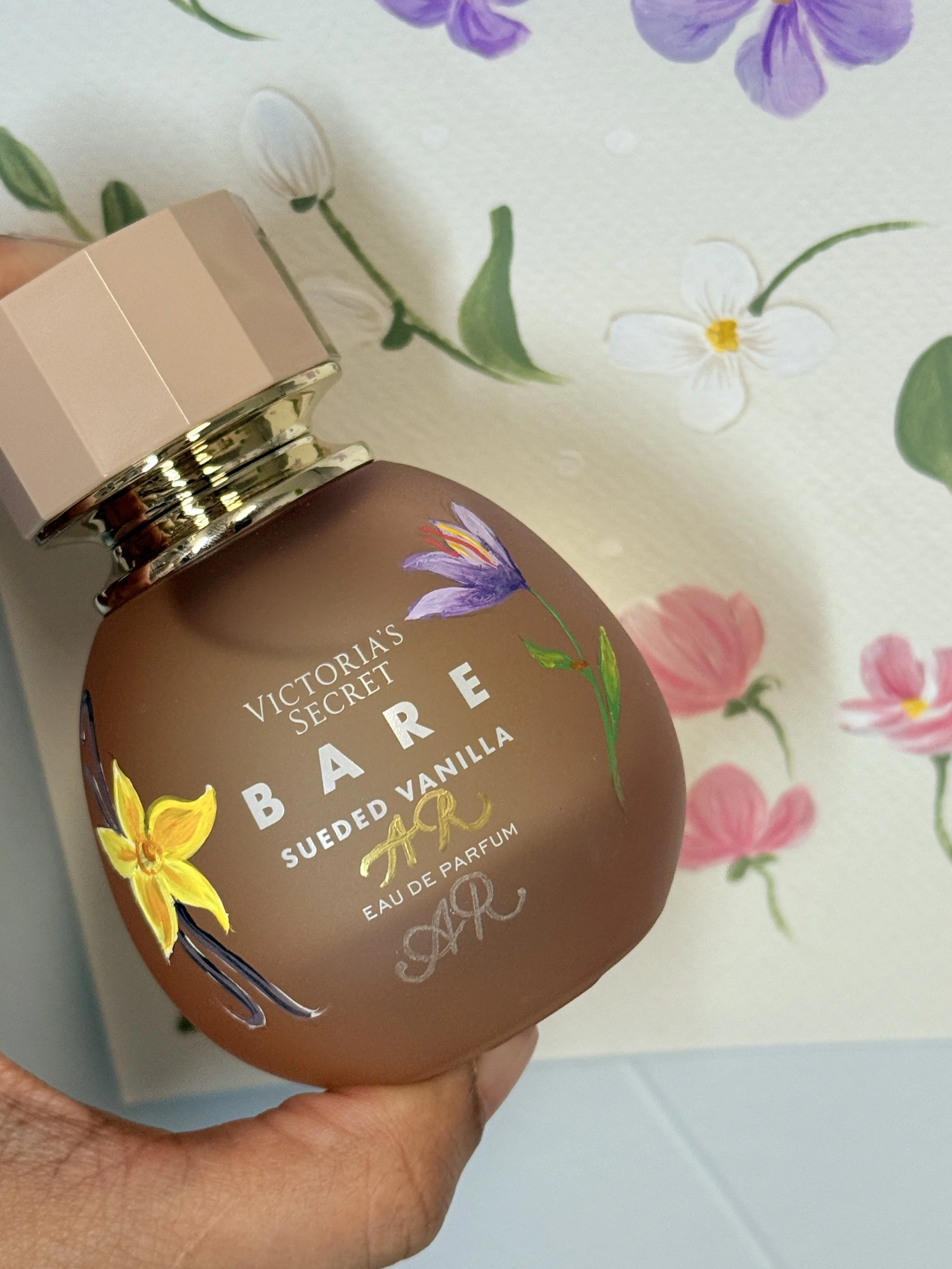 Close-up of a Victoria's Secret Bare Eau de Parfum bottle with a floral label, held in a person's hand against a floral wallpaper background.