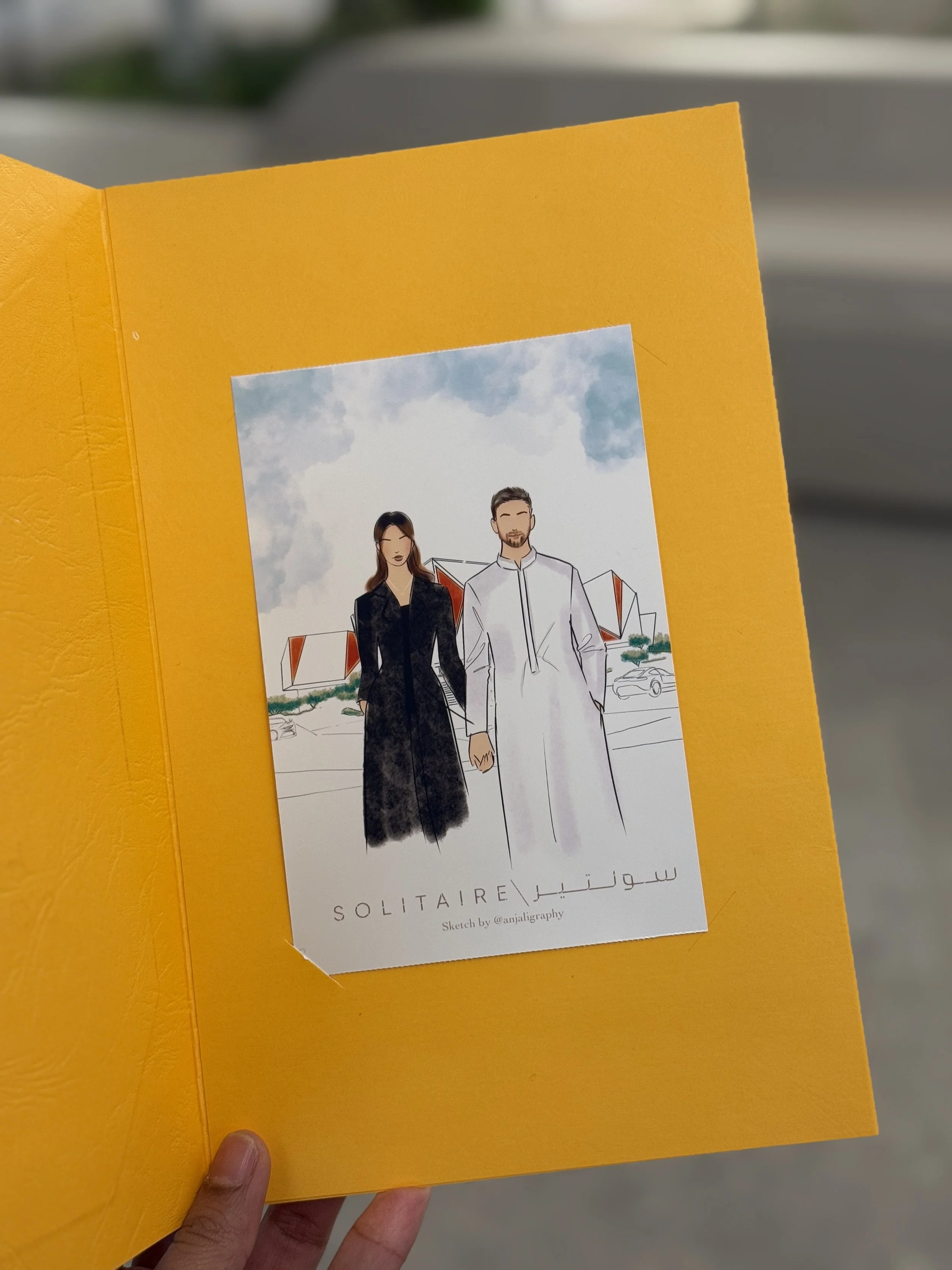 A daytime outdoor scene of a hand holding an orange booklet with a sketch of a man and woman outdoors with buildings in the background, and text "SOLITAIRE" and Arabic text below.