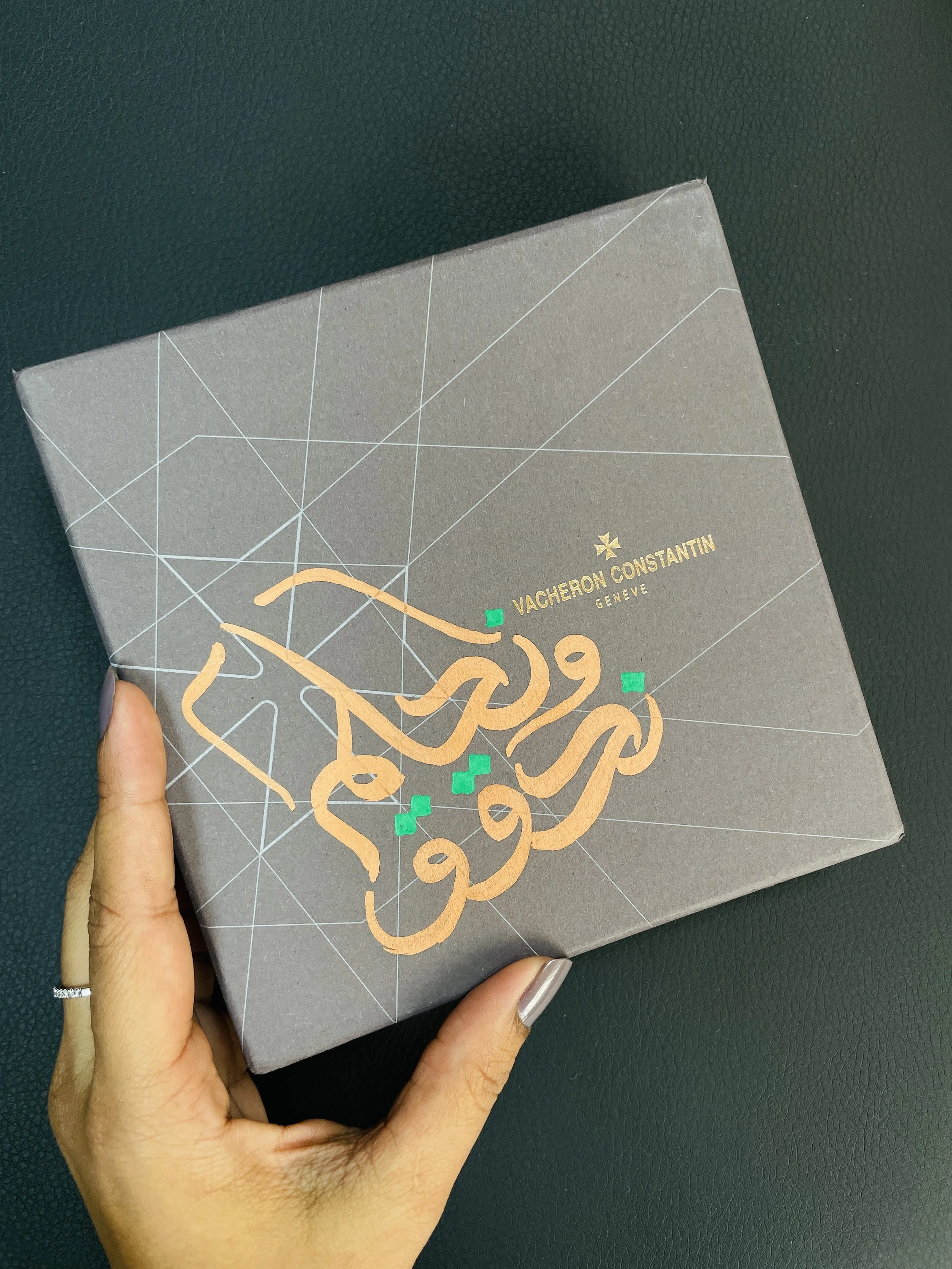 Close-up of a hand holding a gray paper bag with geometric white lines and the Vacheron Constantin logo in gold, and a black textured background.