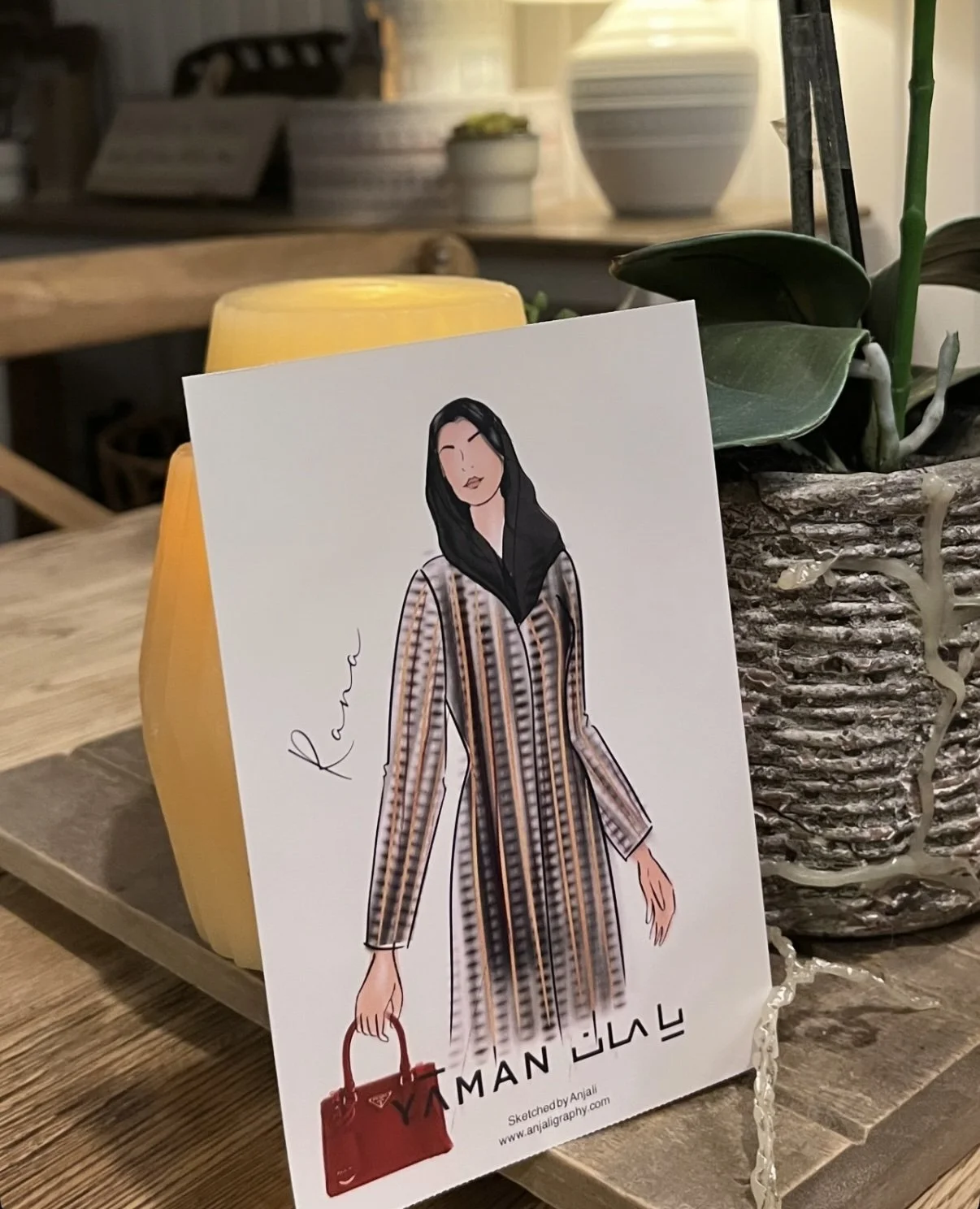 A fashion illustration of a woman with black hair wearing a long patterned coat, holding a red handbag, with the word 'Kane' written on the side, placed on a wooden table next to a yellow candle and a potted plant.