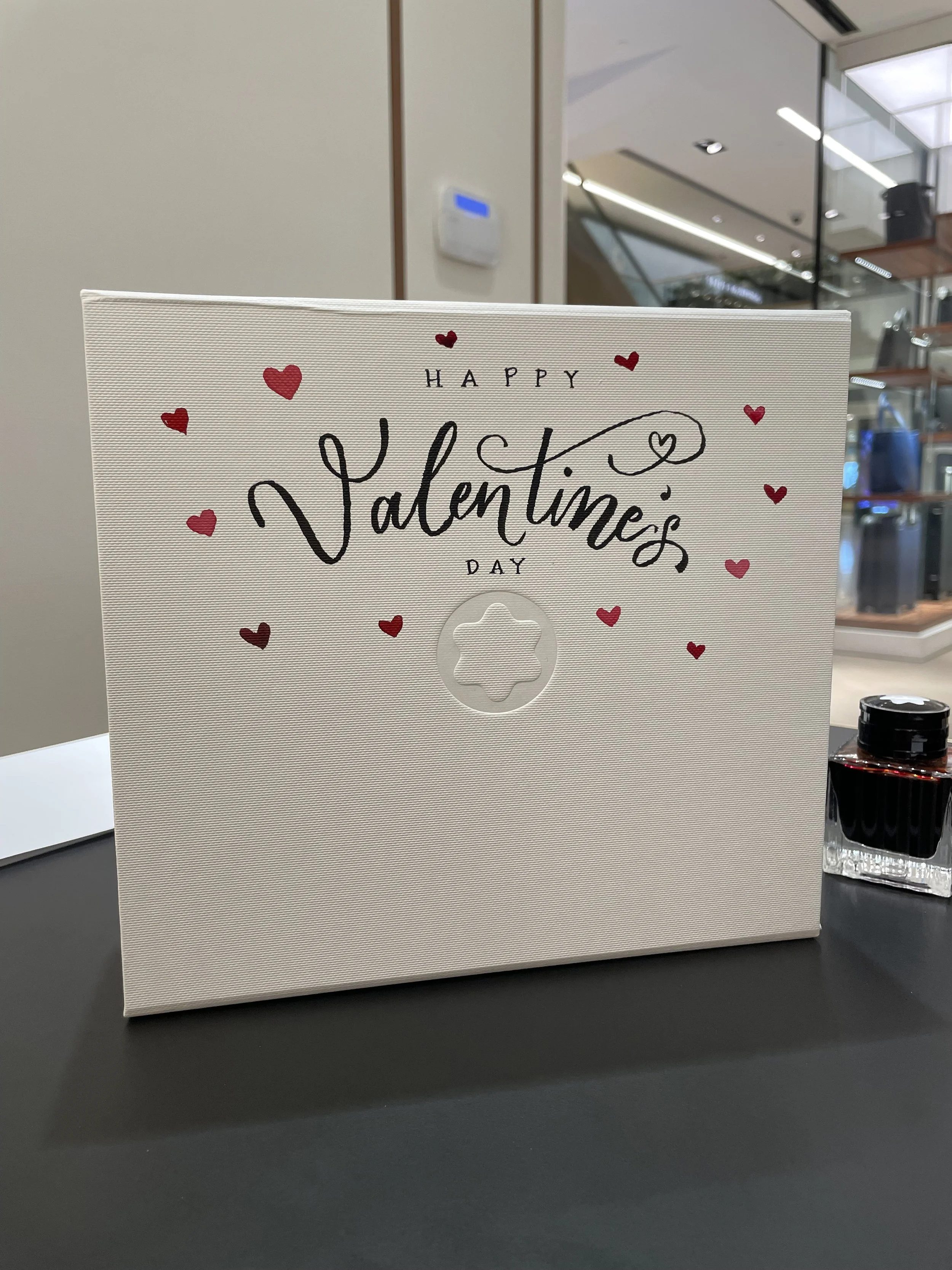 A white Valentine's Day card with red hearts and the message 'Happy Valentine's Day' on a dark surface, with a bottle of ink or lipstick nearby.