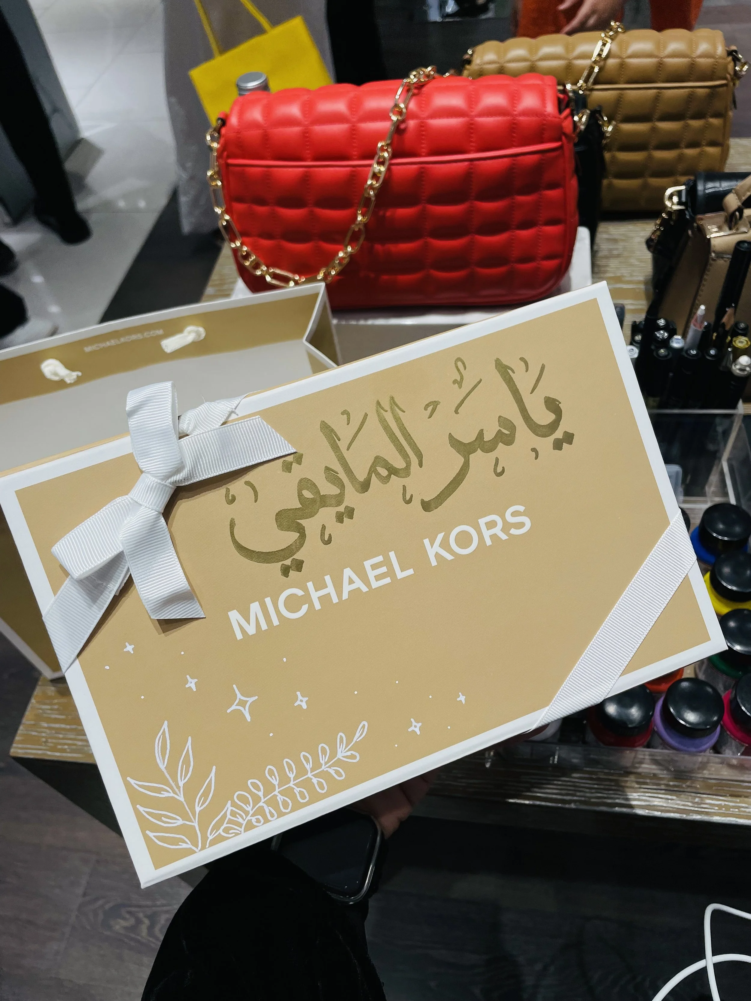 Two Michael Kors branded shopping bags, one gold and one beige, are held by a person in dark clothing, with a display of handbags in the background.