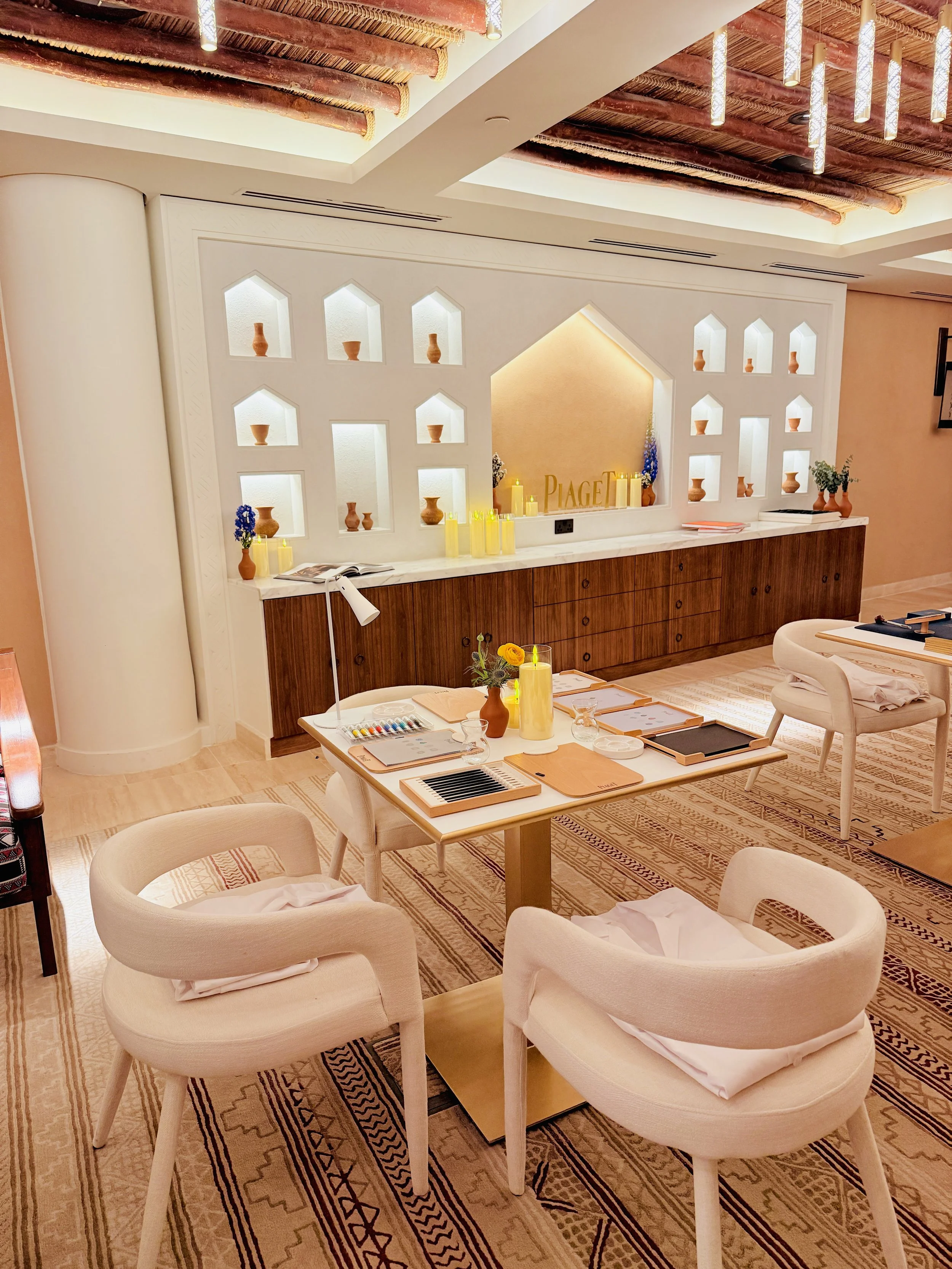 A cozy restaurant or cafe with round cream-colored chairs, wooden tables, and a decorative wall with small vases and candles. The decor includes patterned rugs and a warm, inviting ambiance with soft lighting.