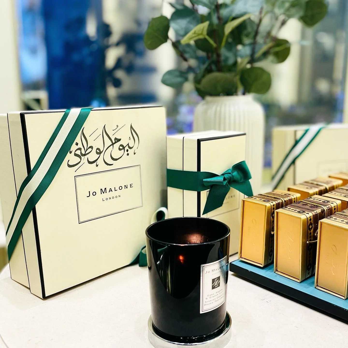 A display of Jo Malone London gift sets, including boxed chocolates with gold wrapping, a black candle with a label, and a smaller boxed item tied with a green ribbon. In the background, there's a potted plant with broad green leaves.
